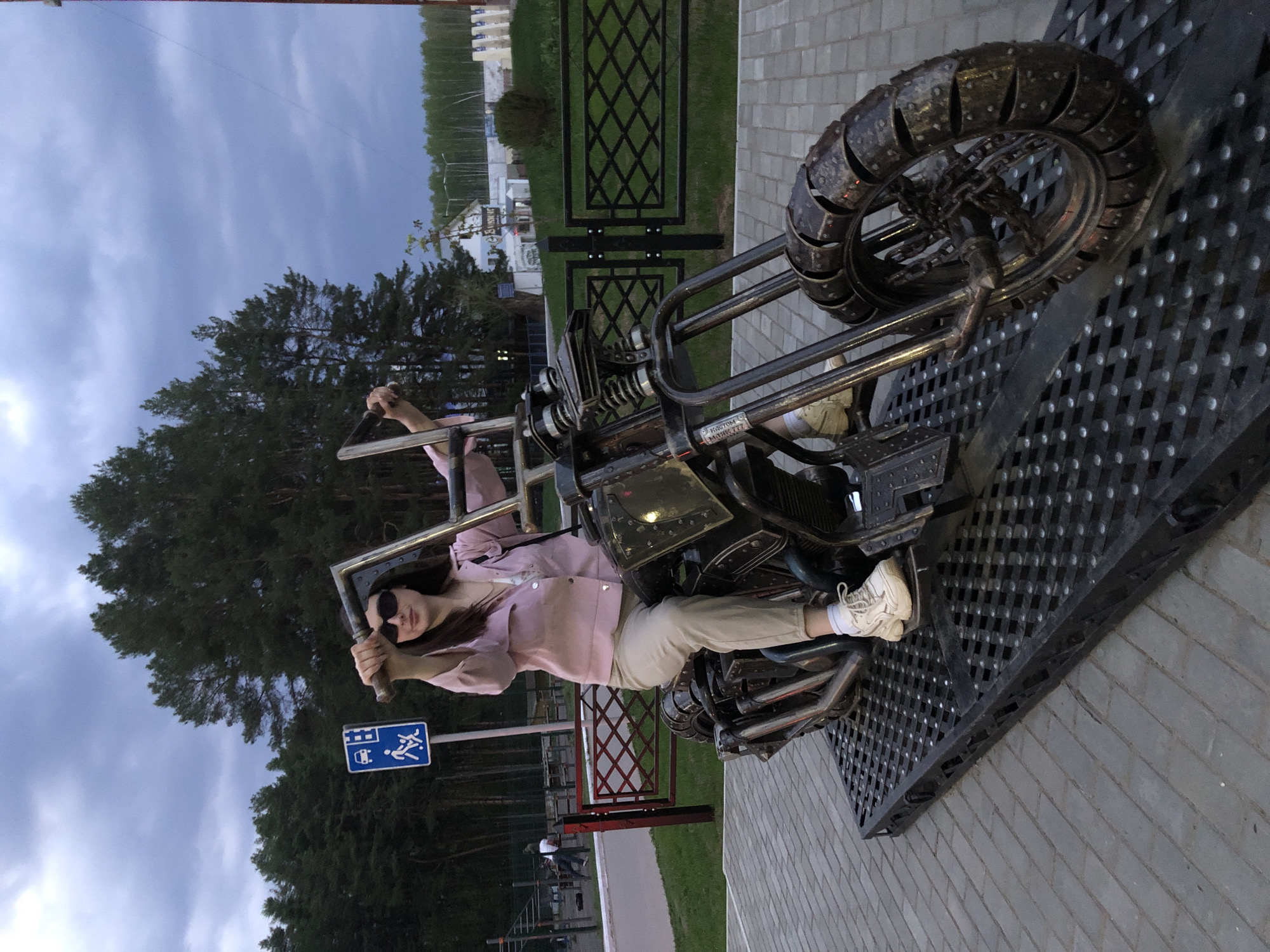 The photo depicts a woman sitting on a metal sculpture of a motorcycle in Chaikovsky, Russia. The sculpture is placed on a gray brick-paved area, possibly a park or public space. Behind her is a wooded area and a blue sign with white pictograms. In the background, a body of water is visible, with buildings and trees along its shore. The sky is overcast. The woman is wearing sunglasses, a pink shirt, and light-colored pants. She is gripping the handlebars of the motorcycle sculpture. A black metal fence is also visible in the background.