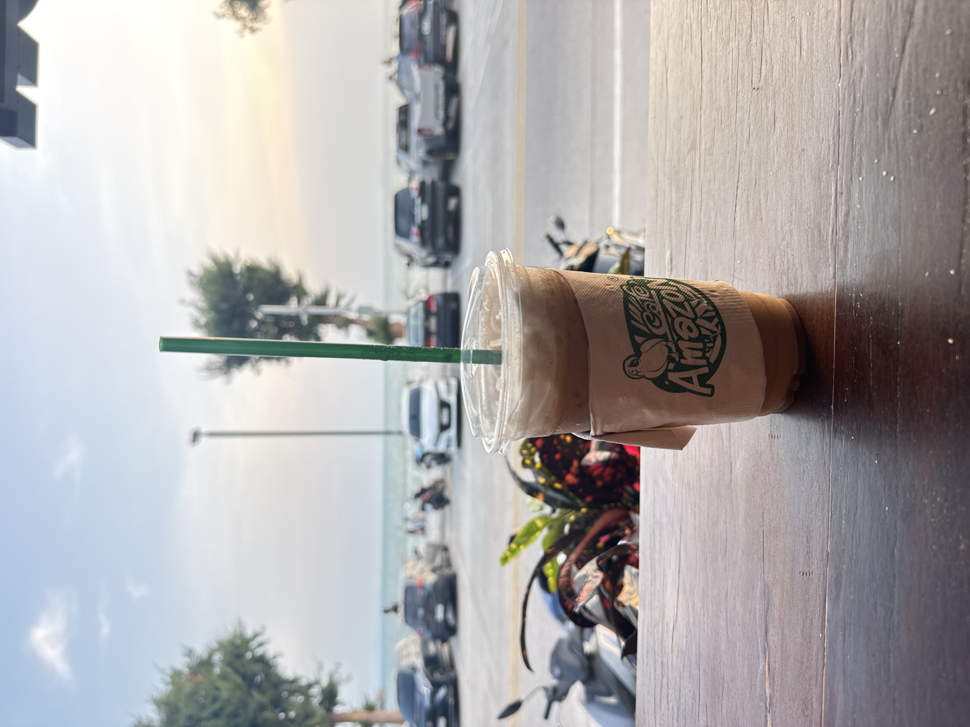 The image, rotated sideways, captures an iced coffee drink placed on a wooden table in the foreground. The clear plastic cup contains a brown beverage and is encased in a beige paper sleeve featuring a distinctive green logo. The logo depicts a monkey or ape-like character and the text "Amazon Cafe" is partially visible. A bright green plastic straw extends from the cup's lid.
The background reveals a bustling coastal street scene under a bright, light blue sky with some soft clouds. A wide road runs parallel to the sea, populated with numerous cars, some parked and others in motion. Beyond the road, the light blue waters of the ocean are visible, extending to the horizon. Patches of green trees and foliage are scattered along the street and in the distance. Streetlights are also discernible. In the mid-ground, colorful potted plants and several parked motorcycles add to the urban tropical environment. The time of day appears to be bright daytime, suggesting late morning or afternoon. The setting conveys a relaxed, outdoor atmosphere, consistent with a cafe or viewpoint in a coastal city like Pattaya, Thailand. No individuals are explicitly seen.