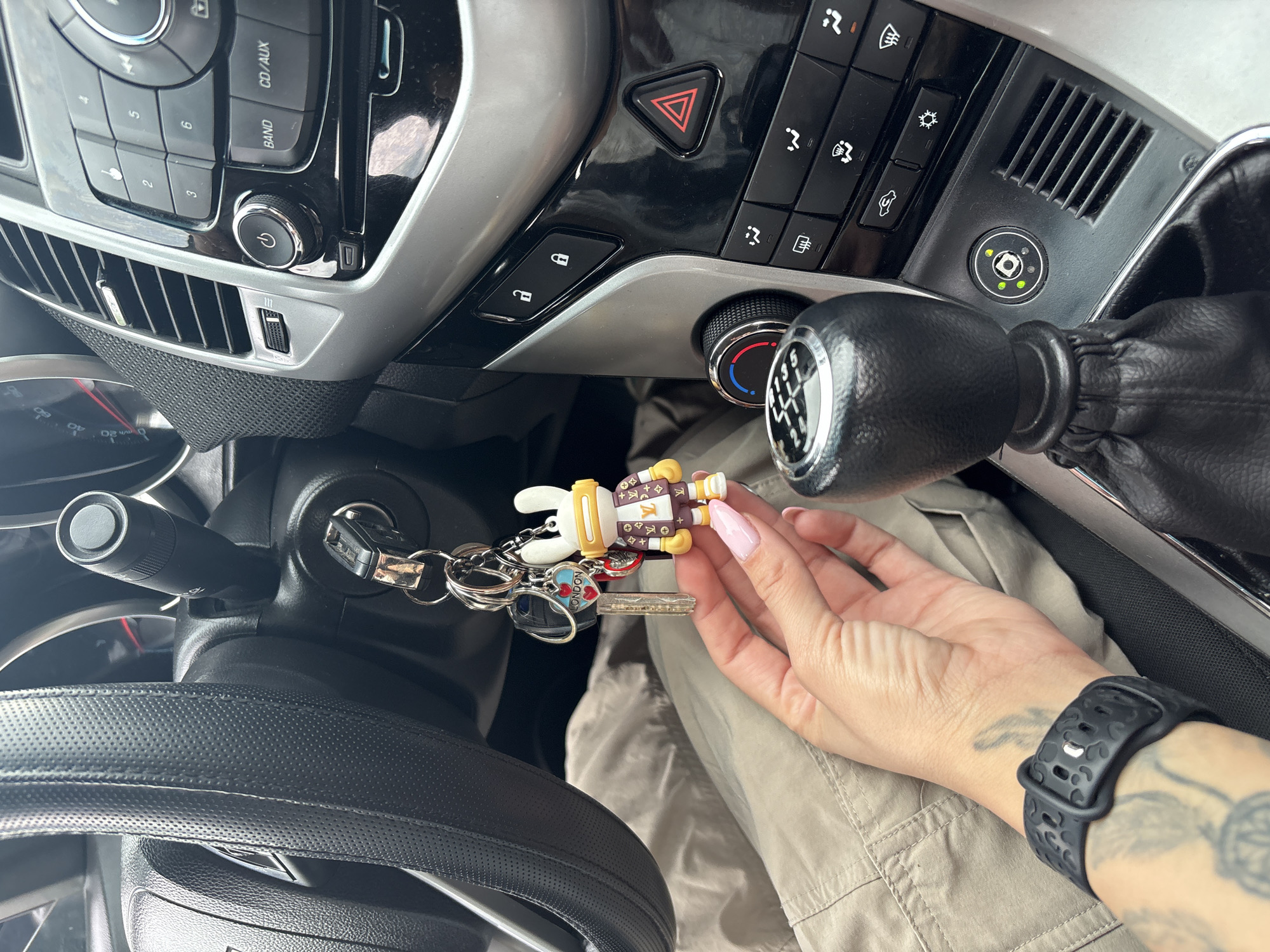 An indoor, slightly rotated photograph captures the interior of a car, focusing on the central console, dashboard, and a person's hand. The setting is a car cabin, likely during the daytime.
The main subjects include a person's hand, characterized by light pink painted fingernails, holding a set of car keys. The key chain features a white, rabbit-like character figurine dressed in a brown and gold suit with "LV" insignias, a heart-shaped charm with "LONDON" inscribed, and other metallic keys. One key is inserted into the ignition on the left, near the steering wheel which has a black textured cover. A black smartwatch with a patterned band is visible on the person's wrist, alongside a faded tattoo on their forearm. The person is wearing light-colored, possibly khaki, pants.
The car's dashboard features a manual gear stick with a black leather boot, displaying numbers 1-6 and R on its knob. The central console includes various car controls: a prominent red triangular hazard light button, door lock and unlock buttons, and climate control dials/buttons. Above these are the audio system controls with buttons labeled "BAND," "CD/AUX," and numbers 1-6, along with a power button on a rotary dial. Below the main console, air vents are visible. A notable circular button with a central "D" symbol surrounded by small green and yellow indicator lights, likely an aftermarket LPG (Liquefied Petroleum Gas) system switch, is positioned near the gear stick. The car appears to be left-hand drive, consistent with the location context of Plovdiv, Bulgaria.