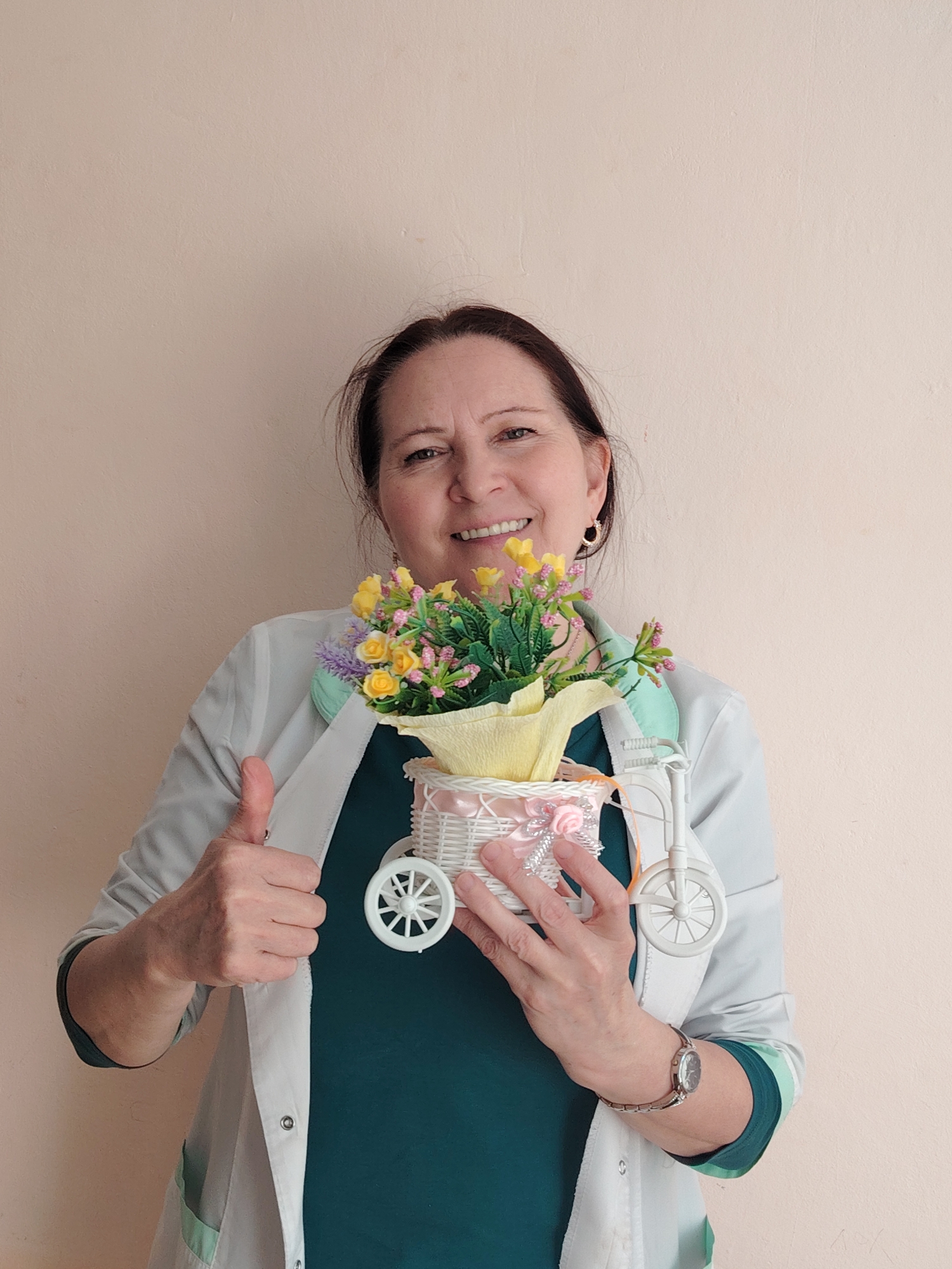 A middle-aged woman with dark hair pulled back and wearing a white medical coat over a dark green top is smiling and holding a small, white tricycle basket filled with yellow and purple flowers. She is giving a thumbs up with her left hand. She is wearing a watch on her left wrist and hoop earrings. The background is a plain, light peach or beige wall. The location is Sarapul, Russia.