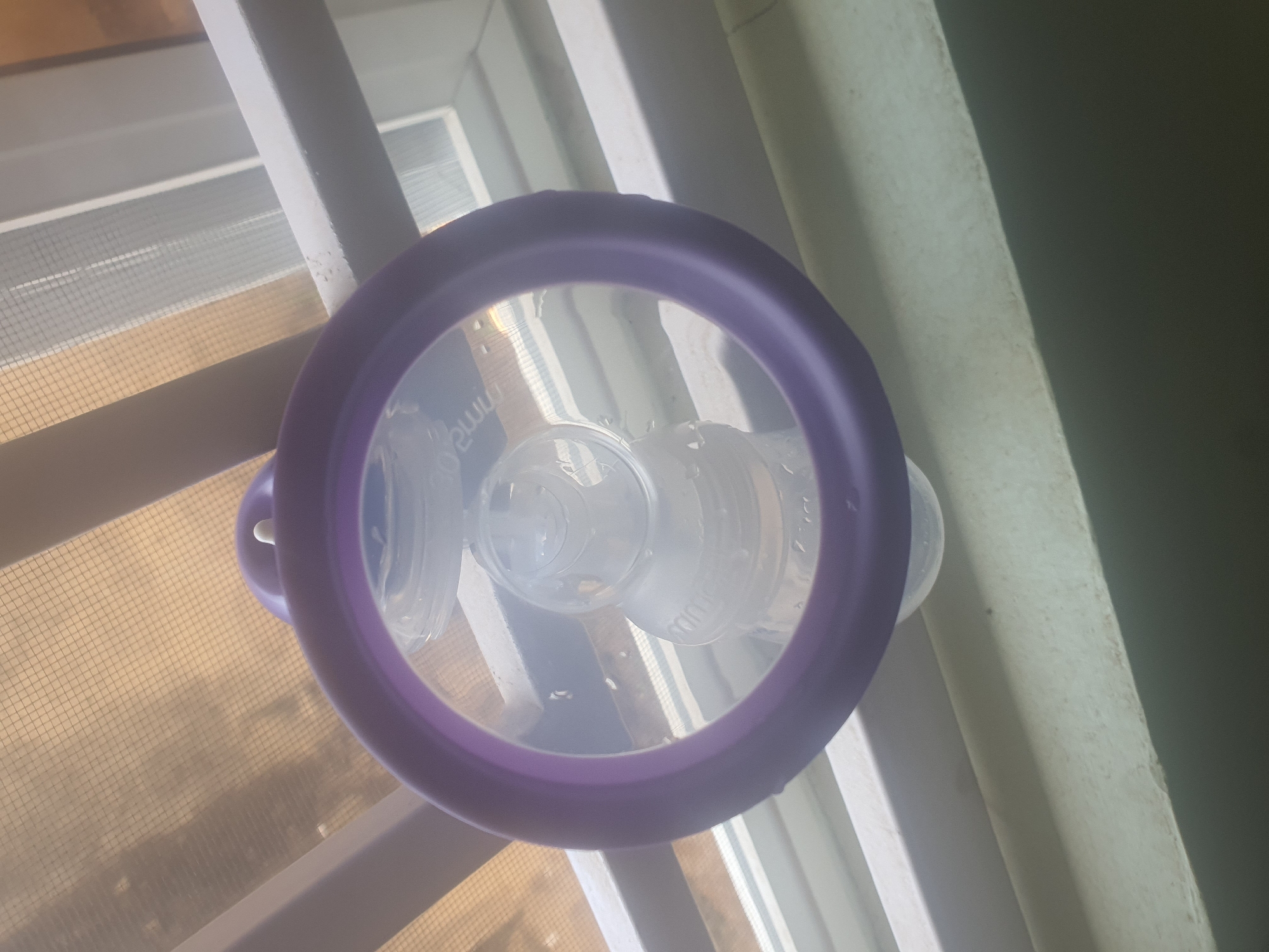 A clear plastic baby bottle with a purple ring around its neck is suspended from a ceiling fixture. The bottle is clear and shows some liquid inside, as well as condensation on its exterior. The ceiling appears to be a textured white material, possibly a screen or textured paint, with horizontal white beams or trim visible. The lighting suggests an indoor setting, possibly a home or a business. The image is taken from a low angle, looking upwards. There are no people visible in the image. The scene is static and offers no clues about the location in Jos, Nigeria.
