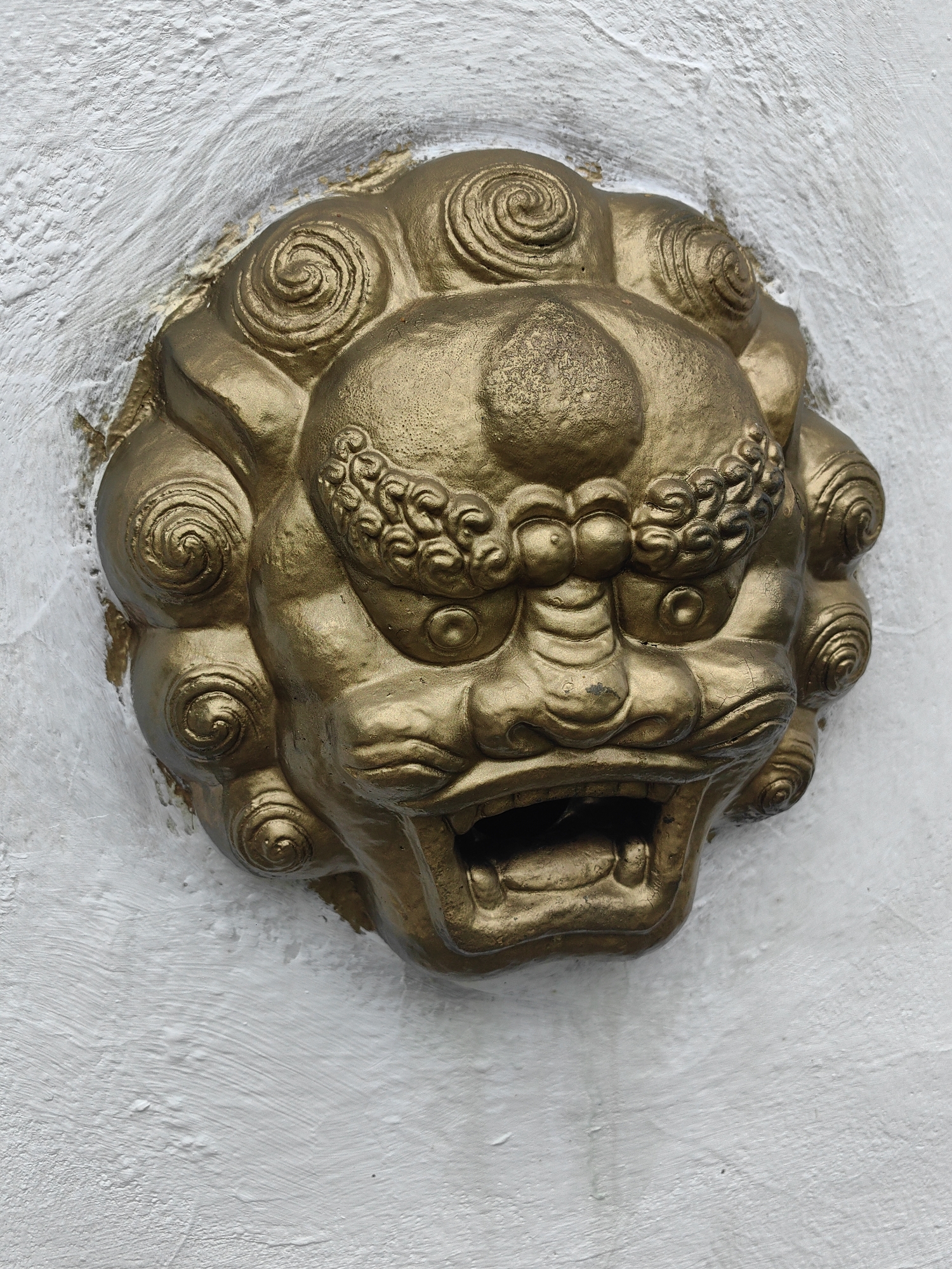 This is a close-up, frontal, eye-level shot of a golden, grotesque lion-like head sculpture protruding from a textured white wall in Tibelti, Russia. The sculpture is highly detailed, with curly manes, prominent cheekbones, bulging eyes, and an open mouth revealing teeth. The gold finish shows some signs of wear and cracking. The wall itself has a rough, painted texture with visible brush strokes. The lighting is even, suggesting an overcast day or an indoor setting. There are no people or animals visible in the image.