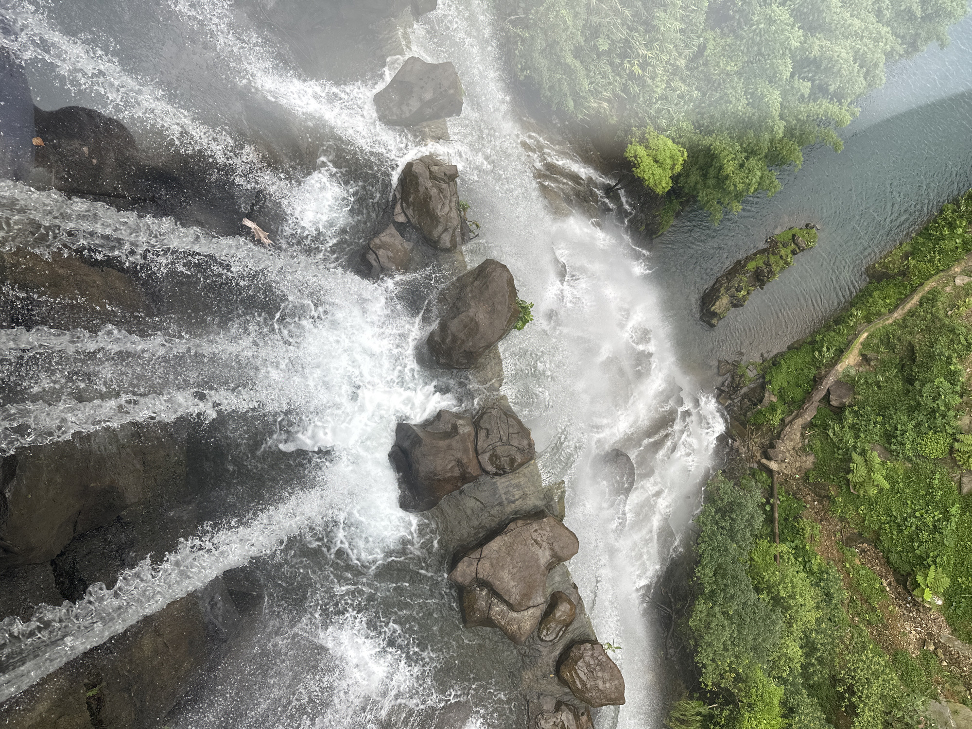 The image is a high-angle, top-down view of a waterfall cascading over rocks into a river. The water is white and frothy, with some of the rocks visible beneath the surface. The riverbanks are lined with lush green vegetation, including trees and bushes. The scene appears to be in a natural, outdoor environment, likely a park or nature reserve. The waterfall is located in Qingyuan City, China. There are no people visible in the image. The image is cropped from the original to provide closer views of different sections of the waterfall and its surroundings.