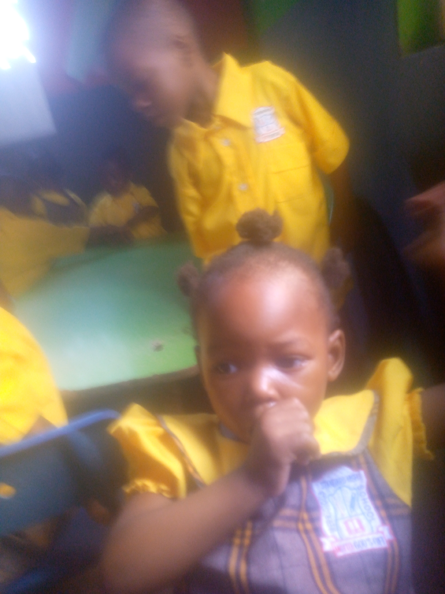 The image shows two young children in what appears to be a classroom setting, presumably in Ikorodu, Nigeria. The foreground child, a young girl with pigtails, is wearing a school uniform consisting of a plaid top with a yellow collar. She has her fingers in her mouth. Behind her, another child, possibly a boy, is wearing a yellow school shirt with a crest. Several other children in yellow shirts are visible in the background seated at a green table. The scene appears to be indoors.
