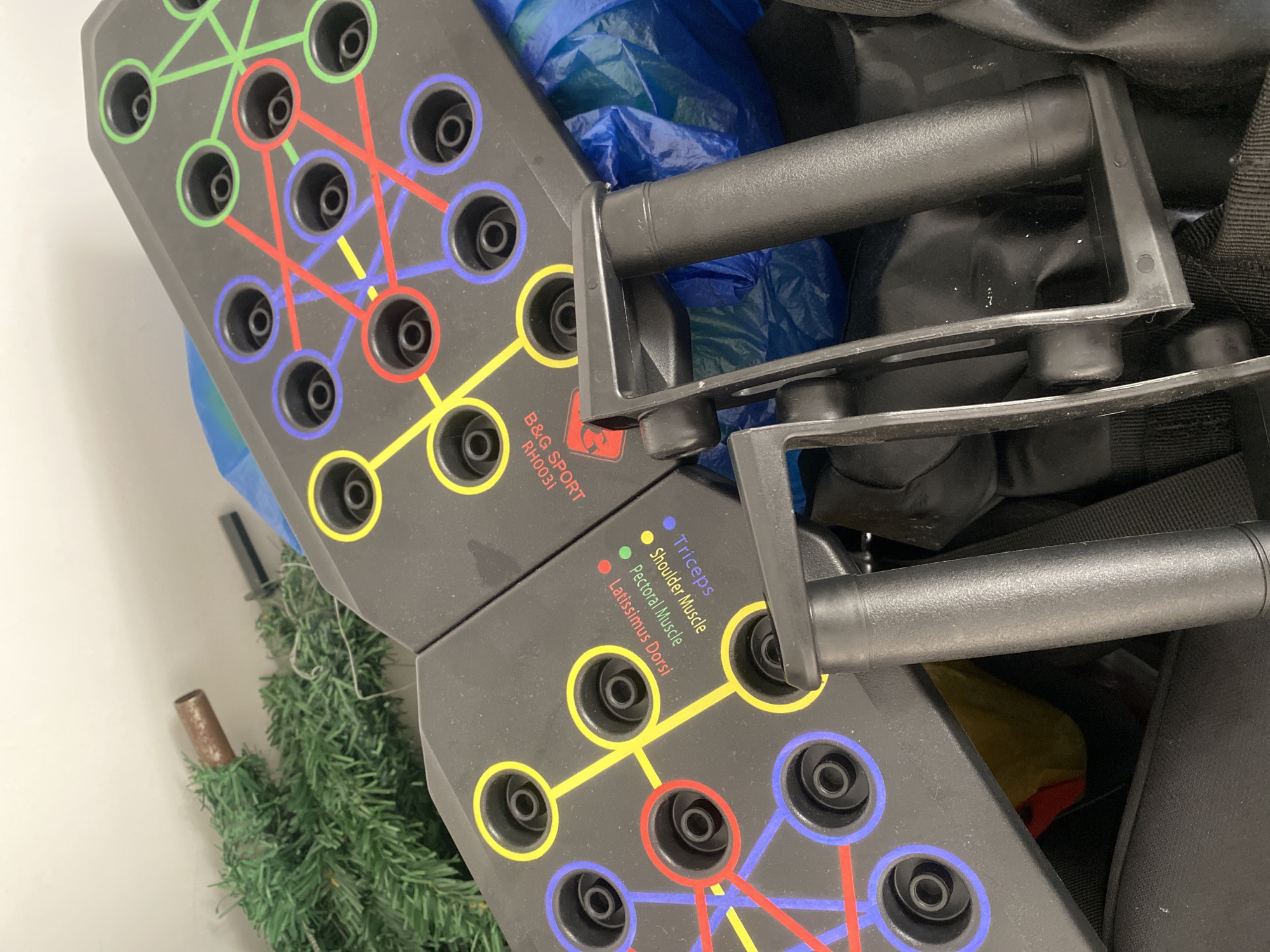 The image shows a set of push-up handles and two gray platforms with labeled circles (Triceps, Shoulder Muscle, Pectoral Muscle, Latissimus Dorsi) connected by colored lines. The handles are black and have a plastic frame. A small, artificial Christmas tree is visible in the lower-left corner. The background includes a blue plastic bag, and a black bag. The scene's location is unknown, and there's no indication it's in Davao City, Philippines. The focus is on the exercise equipment and its arrangement within the frame.