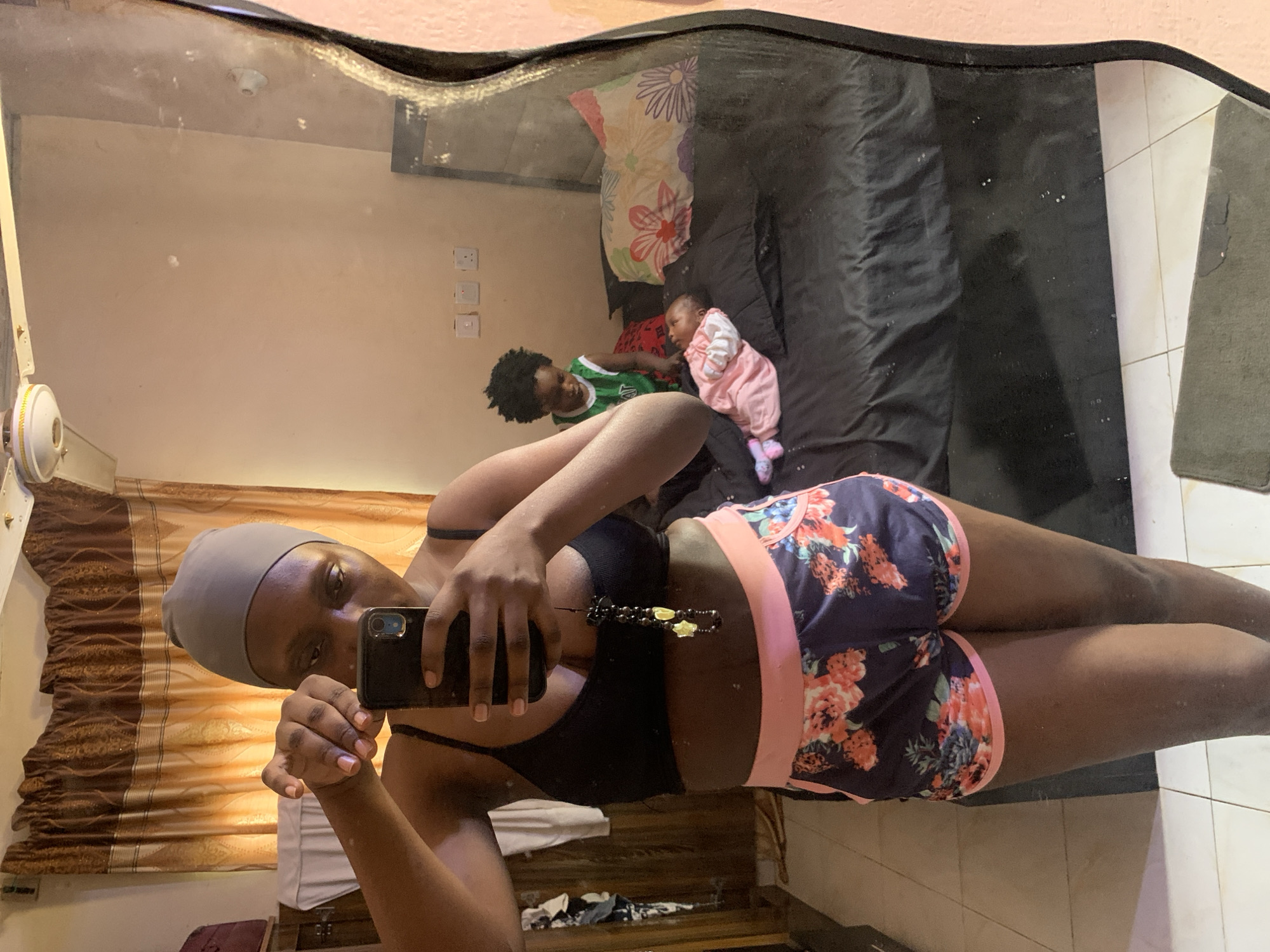 A person takes a selfie in a mirror reflecting a room. In the mirror's reflection, there's an individual wearing a green jersey and a baby lying on a bed with a dark sheet. The bed has a patterned pillow. The room appears to be in a residential setting. The photograph's location is Rafin Bauna, Nigeria, as specified.