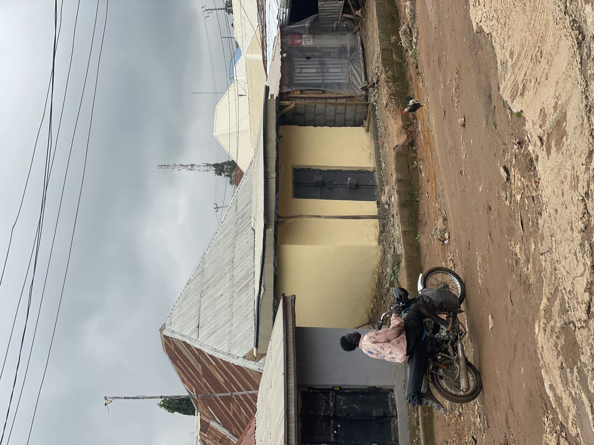 In Kurunduma, Nigeria, a person rides a motorcycle on a dirt road under an overcast sky. The motorcycle is black with a silver fender and a black seat. The rider wears a light pink shirt with a pattern and dark pants. To the left of the road, a grey building has a black door. Next to it is a yellow building with a black door. Behind these buildings are houses with corrugated metal roofs, some appearing rusty. Further in the background, a tall red and white communications tower is visible, along with other buildings and power lines. A rooster is walking on the dirt road in the lower right portion of the image. The environment appears somewhat unkempt, with loose gravel and debris on the road.