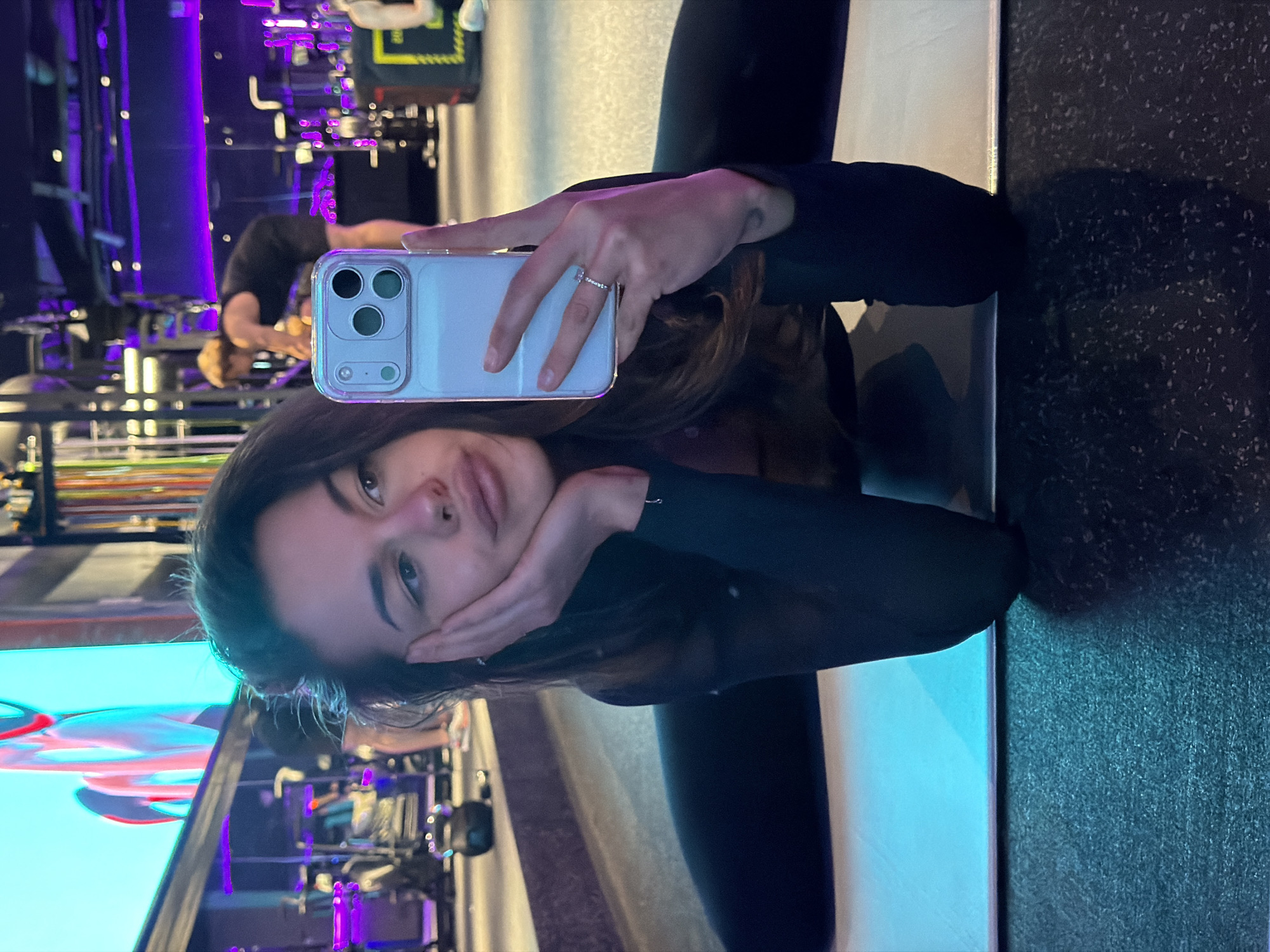 A woman is holding a phone in her hand and taking a selfie in a mirrored room. The woman has long brown hair. She is wearing a black shirt and pants. She has her left hand on her face. The room is brightly lit with blue and purple lights. There are some exercise machines in the background. The floor is dark gray. There are no identifiable landmarks to identify the location in Krasnoyarsk, Russia.