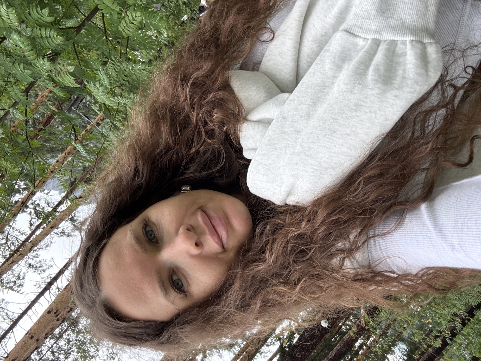A selfie is taken by a woman with long, wavy brown hair and green eyes, looking towards the camera. She is wearing a white ribbed top and a light gray hooded sweatshirt. She has light makeup on, including lipstick and eyeliner. She is positioned outdoors with trees and foliage in the background, suggesting a natural or park setting. The lighting appears to be overcast.
