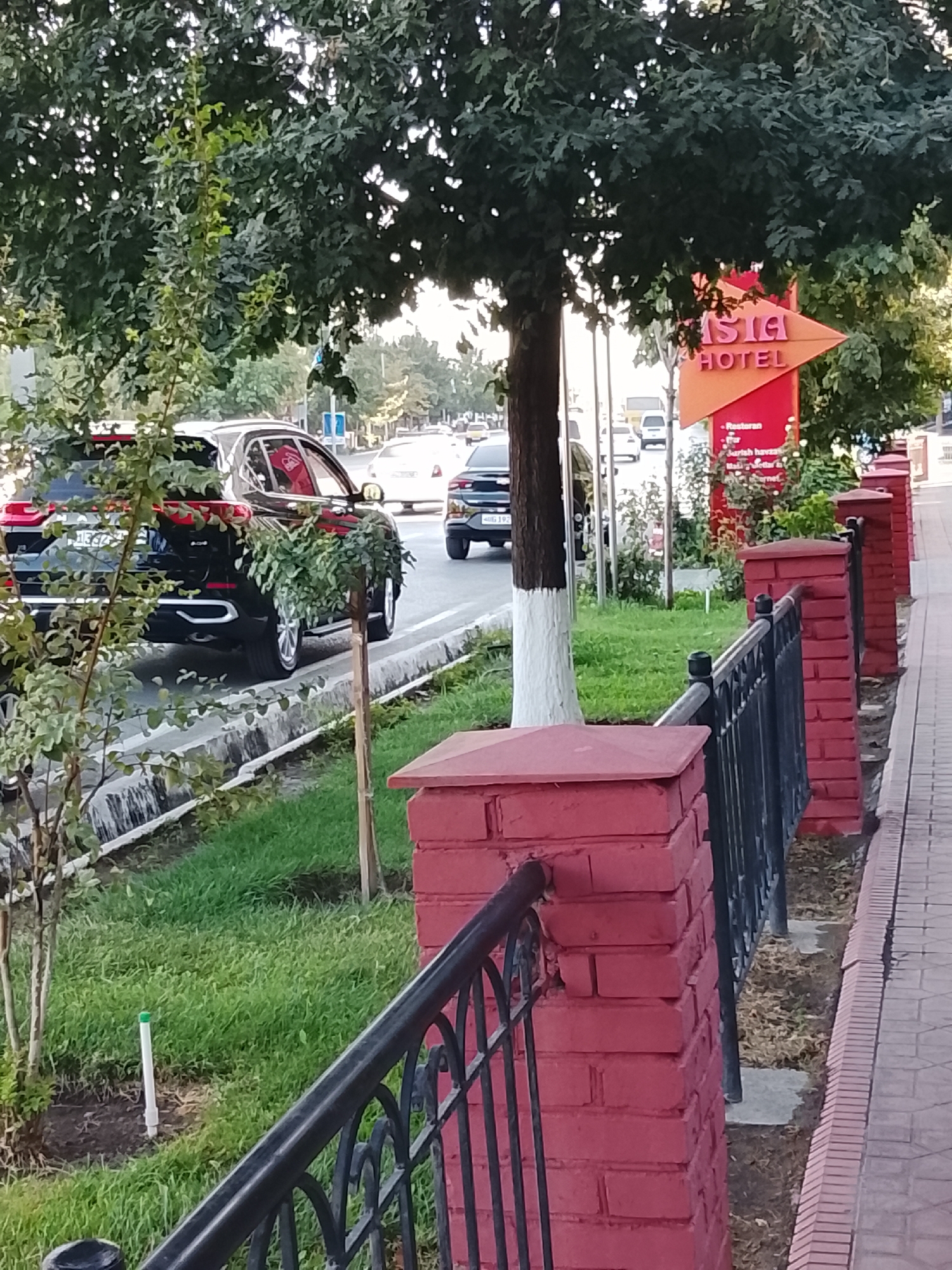 The image shows a street scene in Fergana City, Uzbekistan, with a focus on the "Asia Hotel" sign. The sign is red and shaped like an arrow pointing to the right, with the words "ASIA HOTEL" in white text. The hotel appears to be located behind the sign. In the foreground, there is a red brick fence with black metal railings, and a paved sidewalk. A black SUV is parked on the side of the road, partially obscured by trees. The street is lined with trees and grass, and other vehicles are visible in the background. The weather appears to be sunny, and the time of day seems to be late afternoon or early evening, given the lighting.