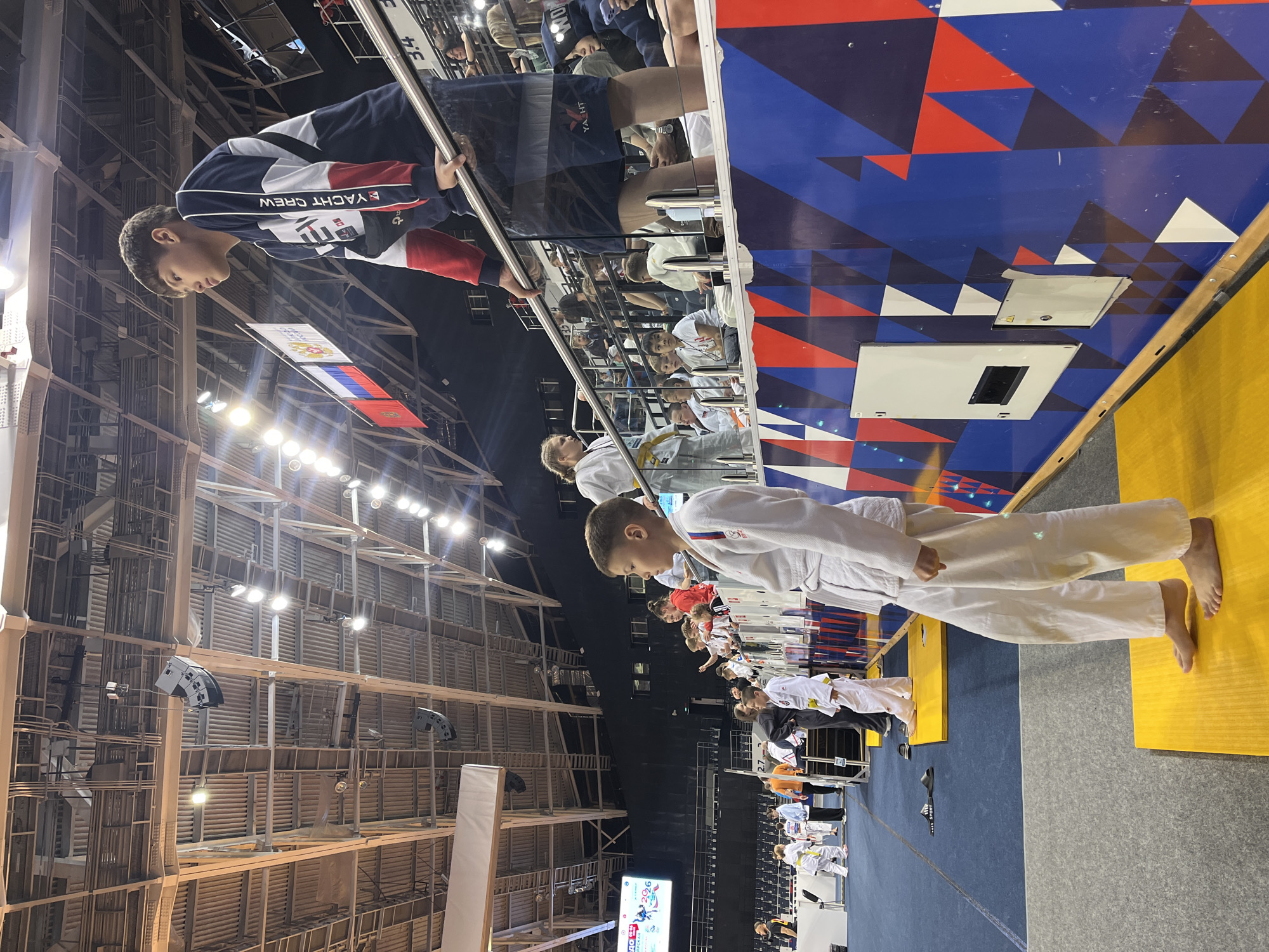 This media file captures an indoor sports event, likely a martial arts competition such as Judo, taking place in a large arena in Krasnoyarsk, Russia.
In the foreground, a young male wearing a blue, white, and red "YACHT CREW" sweatshirt is seen from the chest up, leaning against a metal railing, observing the activity below. His dark hair is neatly cut.
Below the railing, several children and young adults, predominantly male, are dressed in traditional white judogis (martial arts uniforms). Some have yellow belts, indicating their rank. They stand barefoot on bright yellow mats, arranged in lines, suggesting they are waiting for their turn to compete or are being officially presented.
The background reveals a spacious sports hall with a high ceiling characterized by exposed metal scaffolding, beams, and ventilation ducts. Numerous bright spotlights illuminate the area, and large grey speakers are mounted overhead. A Russian flag, featuring the national emblem of a double-headed eagle, is visible in the distance, reinforcing the location context of Russia.
A decorative wall or barrier behind the participants is adorned with a striking geometric pattern of interlocking blue, red, and white triangles. Visible within this pattern are white rectangular access panels. A digital display screen in the far background shows the number "2026" along with some Cyrillic text and event branding. The floor in the activity area is a mix of blue and grey matting. The overall atmosphere suggests a formal sporting competition or demonstration.