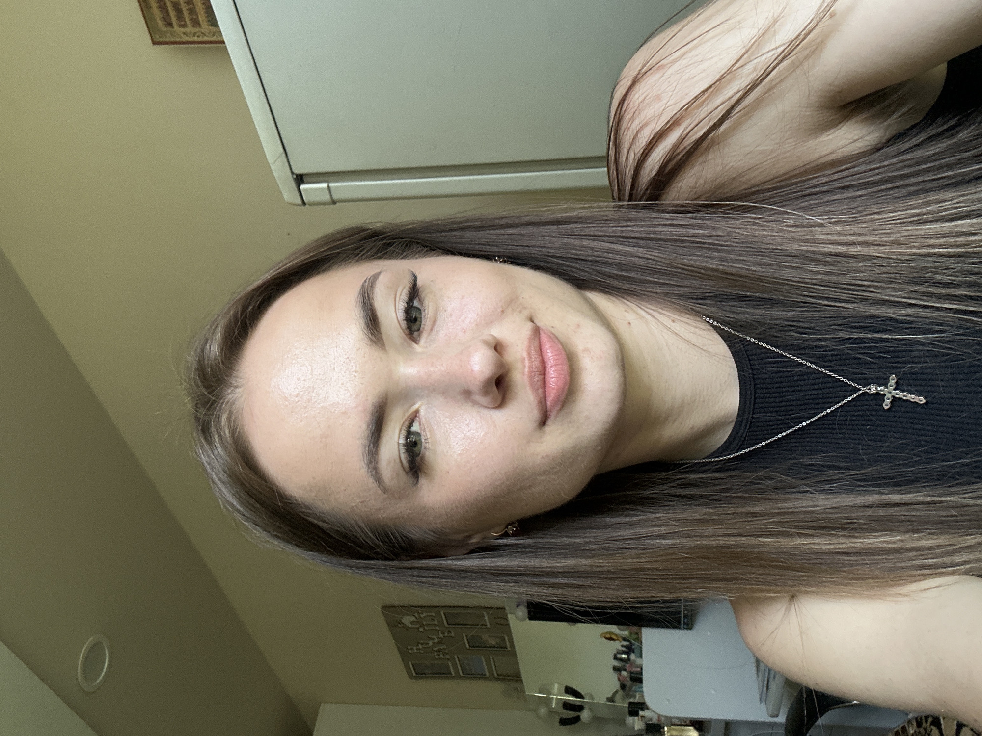 A close-up self-portrait of a female individual with a fair complexion is depicted indoors in the city of Unknown, Unknown. The subject has long, straight brown hair, parted slightly off-center, extending over her shoulders. She possesses light green eyes, dark eyebrows, and visible eyelashes, with subtle eyeliner. Her lips are lightly colored and gently closed, forming a slight upward curve. The individual wears a sleeveless black ribbed top and a silver-colored chain necklace featuring a cross pendant. Small, dark hoop earrings are present in both ears. The background on the left reveals a light-colored wall with a multi-framed picture collage, below which a vanity surface holds various cosmetic bottles and a mirror reflecting blurred objects. On the right, the light-colored wall continues, alongside the upper section of a light-grey refrigerator with a metallic trim. A small framed item is mounted on the wall above the refrigerator. A recessed circular light fixture is visible on the light-colored ceiling in the upper left of the frame. The scene is illuminated by ambient light, highlighting the subject's facial features and the immediate environment.