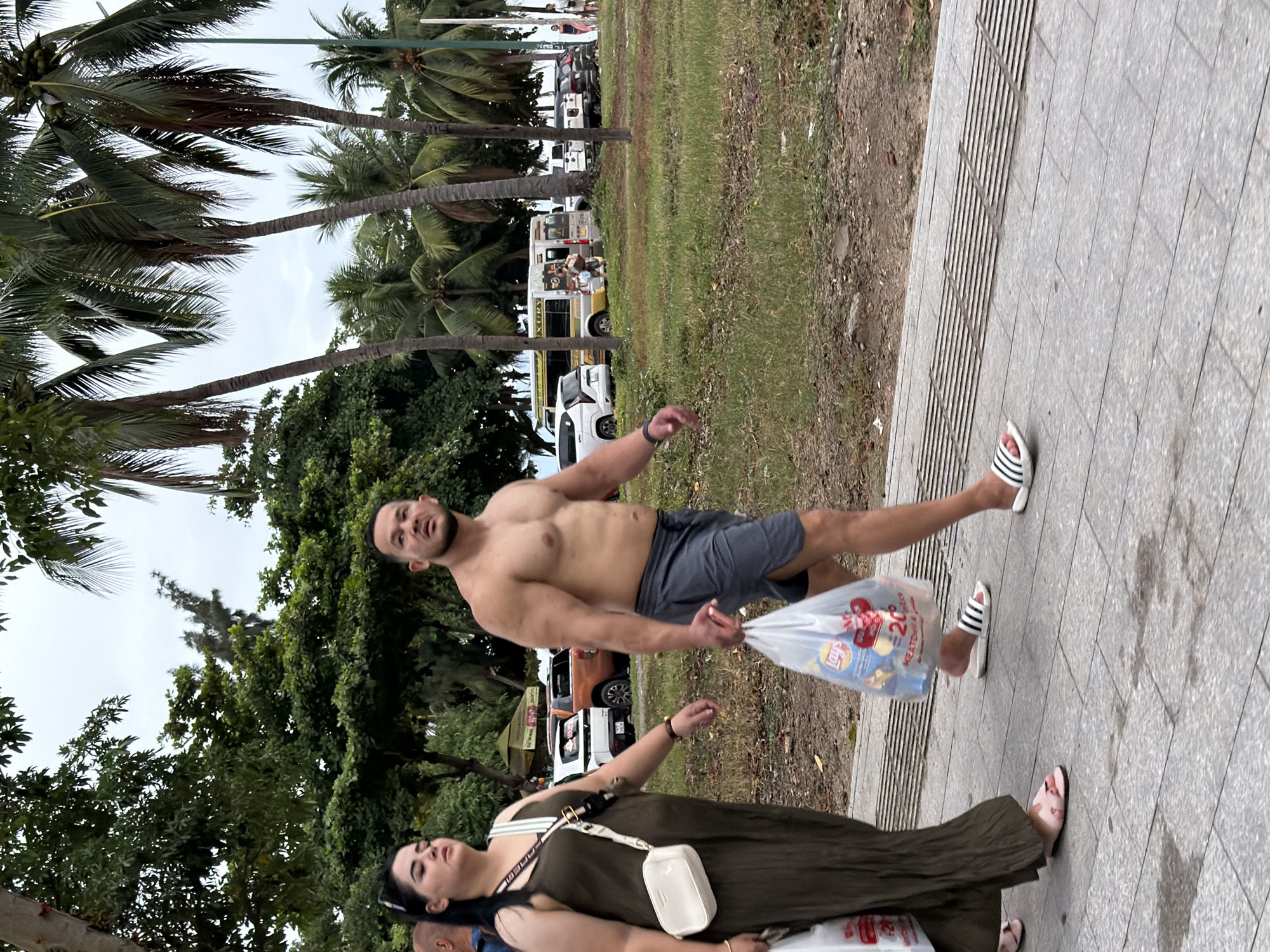 A man and a woman are walking on a paved sidewalk in front of a grassy area with palm trees. The man, who is shirtless, is carrying two plastic bags filled with items, including a bag of Lay's potato chips. He is wearing grey shorts and black and white striped slides. The woman is wearing a long, olive-green dress and a white crossbody bag. She is carrying a white plastic bag in her left hand. In the background, several cars are parked along a road, including a white SUV, an orange pickup truck, and a white van with yellow and black lettering. The sky is overcast. The location is identified as Nha Trang, Vietnam.