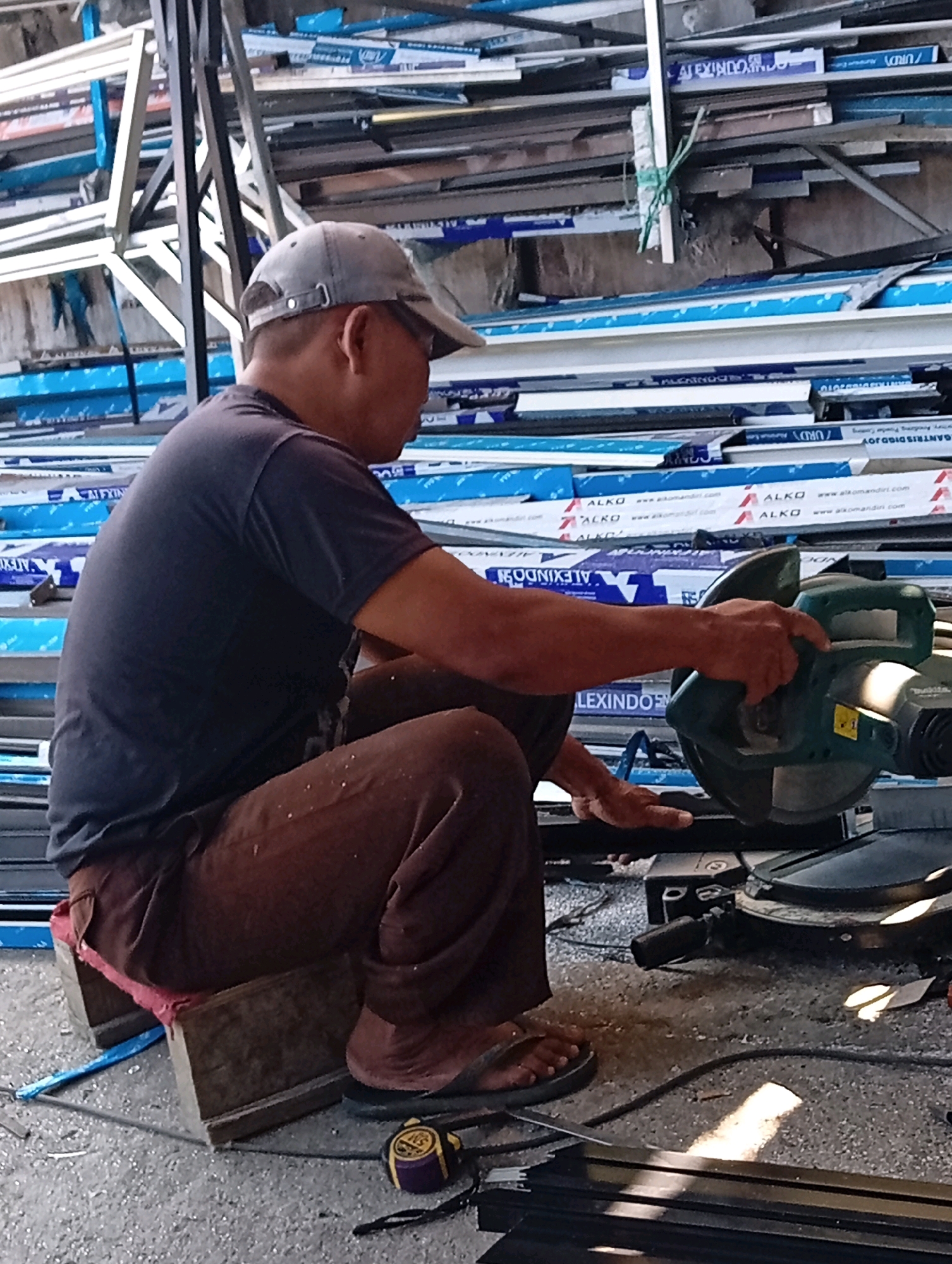 A man, wearing a dark t-shirt, brown pants, a grey baseball cap, and flip-flops, is crouching on a wooden stool in what appears to be a workshop or storage area. He is operating a green and black power saw, likely a miter saw, and is positioned to cut a piece of metal. The background is filled with stacks of aluminum extrusions, some with blue protective film. The floor is littered with metal shavings, indicating recent cutting activity. A yellow and purple measuring tape lies on the ground near the man's feet. The scene suggests industrial or construction-related work.