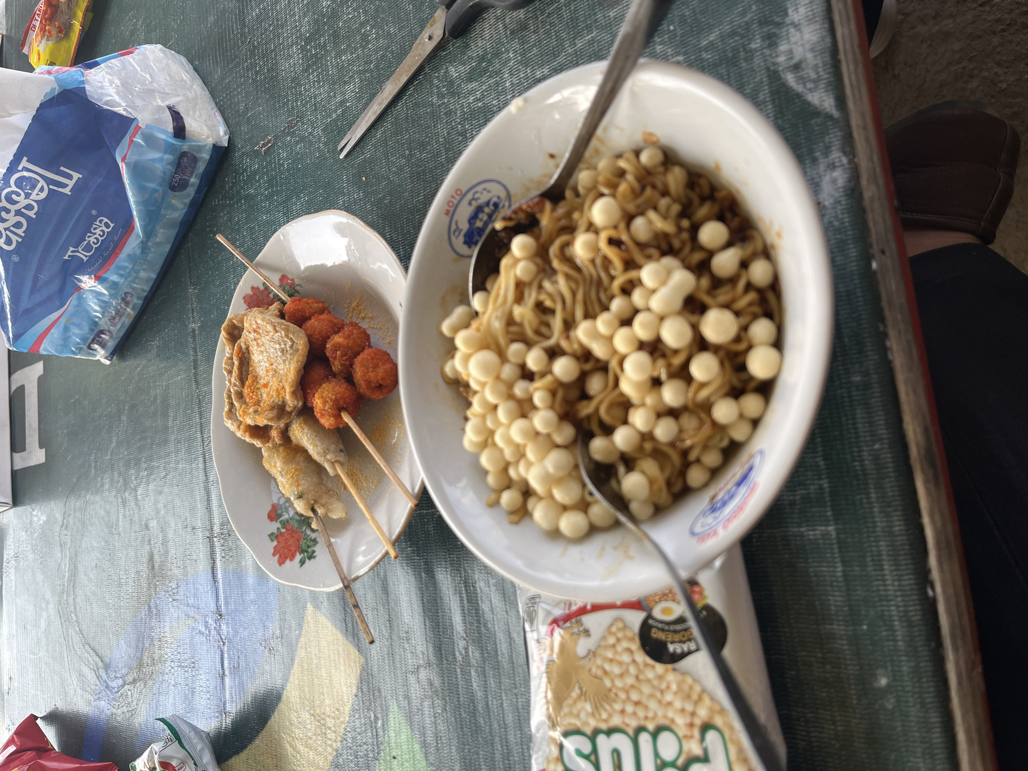 The image depicts a close-up view of a table set for a meal. In the foreground, a white bowl contains a generous portion of instant noodles, topped with numerous small, pale, spherical snacks. A metal spoon rests in the bowl, partially submerged in the noodles. To the left of the bowl is a torn open package of these same spherical snacks. In the background, a white plate holds several skewers of food: some appear to be fried or grilled meat or fish, while others are coated in a reddish-orange spice or batter. A few larger, flatter pieces of food are also on the plate. A pack of "Tessa" brand tissues is visible behind the plate. A pair of scissors lies to the right of the plate. The table surface has a patterned, textured appearance, possibly a tablecloth or placemat, with colors including green, blue, and yellow. The overall lighting suggests an outdoor or well-lit indoor setting. The scene appears to be a casual meal setting, possibly at a street food stall or a small eatery in Gembong, Indonesia, given the presence of Indonesian snacks and the overall atmosphere. No people are directly visible in the frame, but their presence is implied by the prepared food and dining utensils.