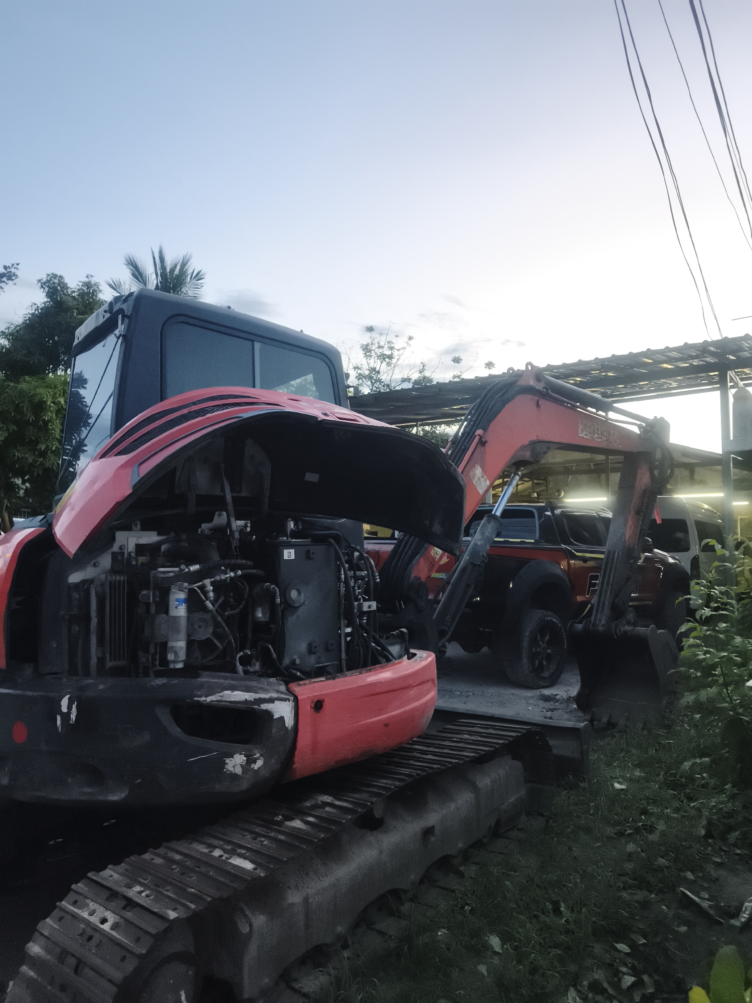 The image shows a red and black Kubota excavator with its engine compartment open. The excavator is parked under a corrugated metal roof in Baan Mae Nam, Thailand. Behind the excavator, there is a red pickup truck and a white van. Green vegetation surrounds the edge of the parking area. The sky is partially visible with light clouds and trees in the background. The excavator's treads are worn.