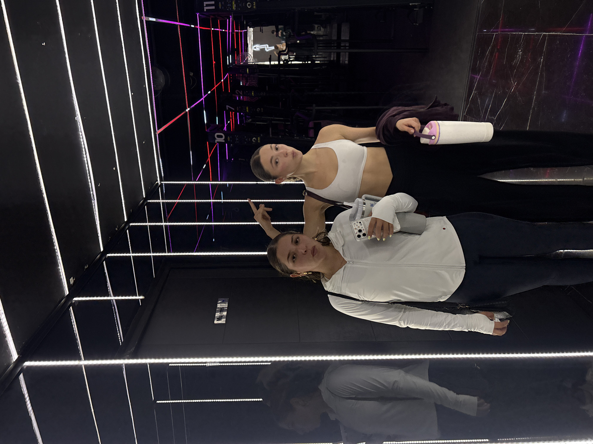 Two young women are posing for a photo in what appears to be a gym. The gym has black walls with bright white and purple neon light strips. Exercise equipment is visible in the background. The women are standing in front of a door.
The first woman is wearing a white sports bra and black leggings, holding a white water bottle. The second woman is wearing a white jacket and black leggings, holding a phone. Both women are looking at the camera.
The sign on the door says "Mujeres".