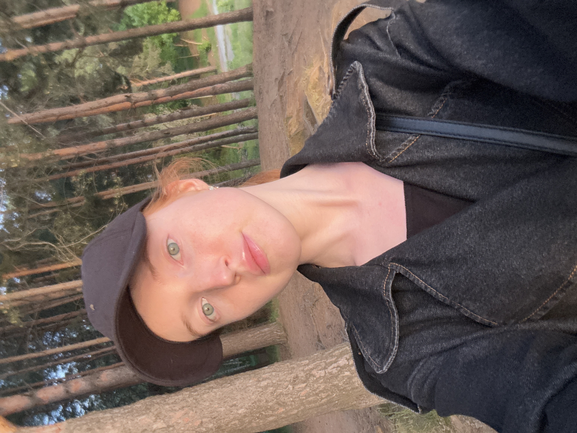 A medium close-up shot depicts a person with fair skin, light red hair, and green eyes wearing a black baseball cap and a black denim jacket with visible seam details. The person is positioned at an angle in the frame, with their face angled towards the camera. A thin black strap of a bag or purse is visible across their shoulder. The background reveals a forest setting, with numerous tall, thin trees with light brown bark standing closely together. The lower portion of the background suggests a grassy or dirt path. Based on the environment and mention in the prompt, the location is likely a forest or park in Chaikovsky, Russia.