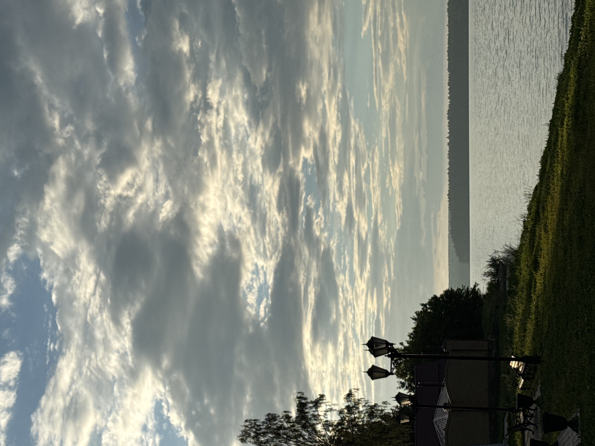 This image depicts a tranquil lakeside scene in Votkinsk, Russia. The sky is filled with a dramatic, cloudy formation, with patches of blue peeking through. In the foreground, a grassy embankment slopes down towards the calm, reflective water of the lake. Several lampposts with ornate designs stand along the embankment, casting long shadows. To the left, a building with a brick facade and a dark-tiled roof is visible, partially obscured by trees. The trees themselves, including a tall, slender birch, add a natural element to the landscape. A distant tree line forms a dark silhouette across the horizon. The overall impression is one of quietude and natural beauty, characteristic of a serene summer day in the Russian countryside.