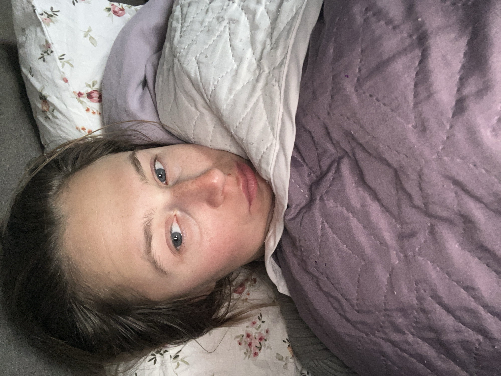 A woman is lying in bed. She has fair skin, light brown hair, and blue eyes. Her head rests on a floral-patterned pillow. She is partially covered by a lavender-colored quilt. The environment appears to be an indoor bedroom setting. The location, Rafailovići, Montenegro, cannot be verified from this image alone.