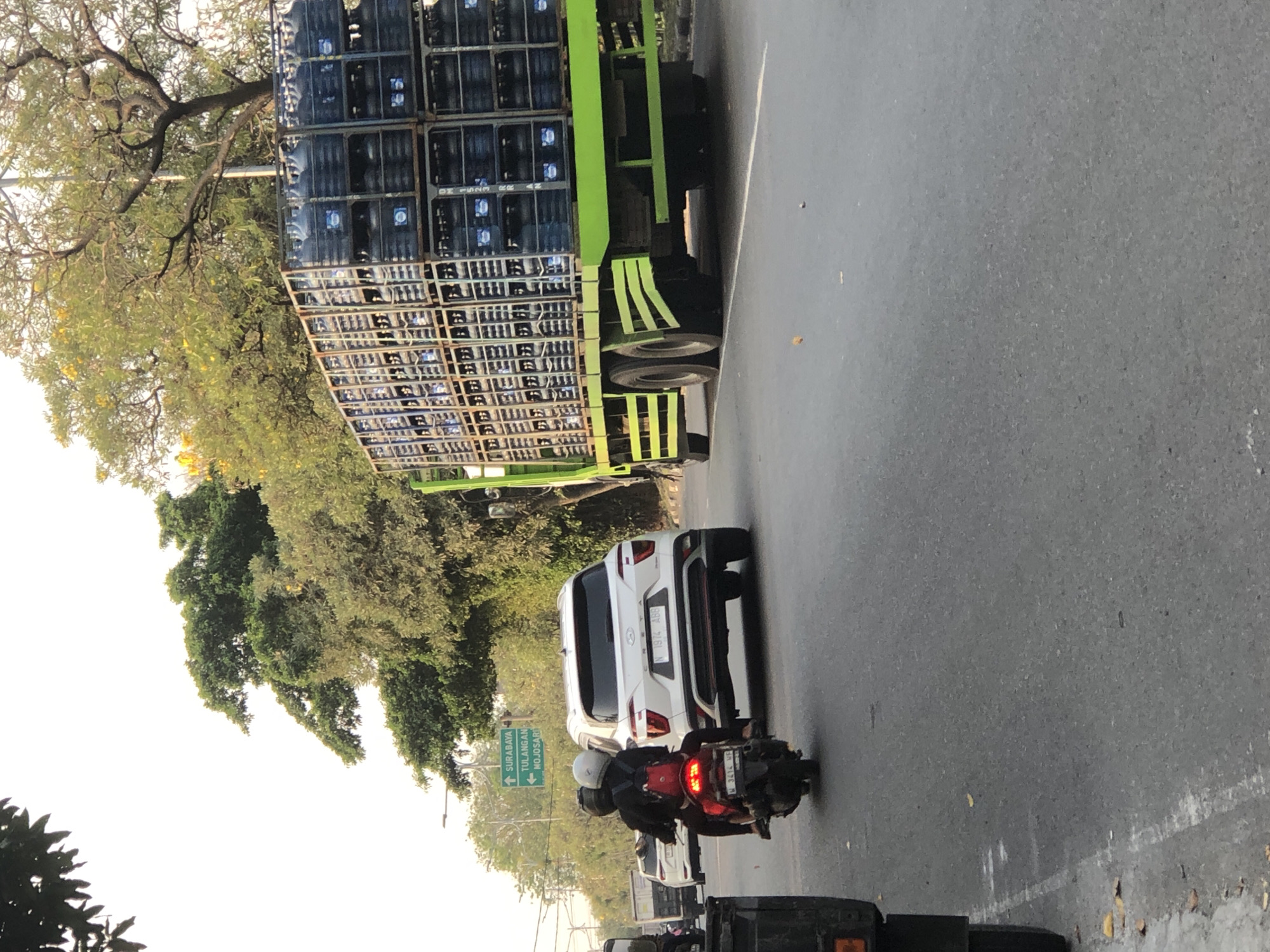 The image captures a street scene in Kludan, Indonesia, likely during the day given the lighting. A large, green truck is prominently featured, loaded with numerous stacked blue water gallon containers in metal racks. The truck's tires and lower chassis are visible. To the left of the truck, a white SUV is driving on the asphalt road. In front of the SUV, a motorcycle with two riders is moving away from the camera. The rider in the back is wearing a red backpack. A street sign with green text indicating "SURABAYA," "TULANGAN," and "MOJOSARI" is visible in the background, along with trees and overhead wires. The road surface is asphalt with some white lane markings and scattered debris. The overall environment suggests a moderately busy road with vehicle traffic.