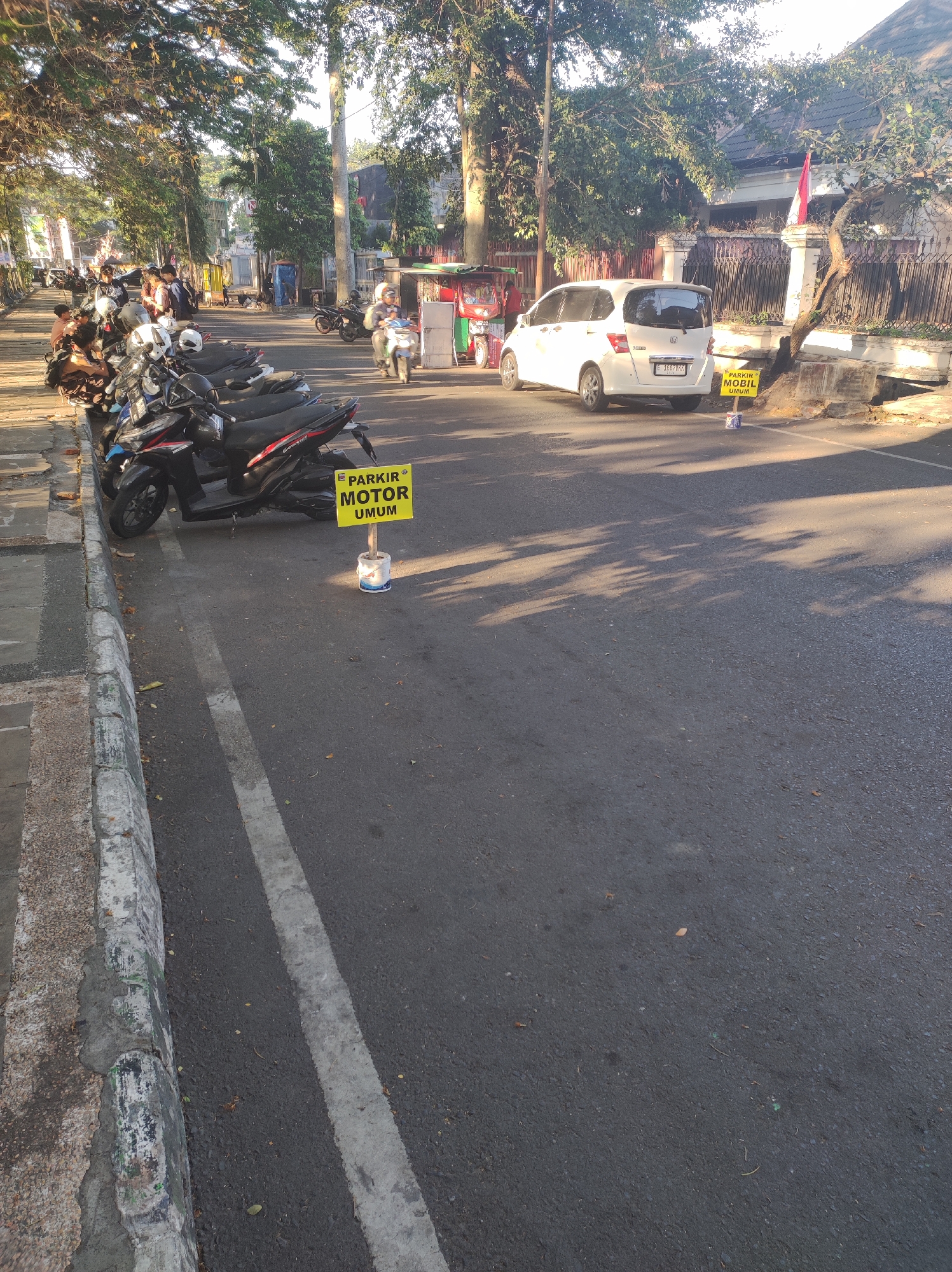 The image depicts a street scene in Malang, Indonesia, likely in the morning given the long shadows. On the left side of the road, a row of parked motorcycles is visible, with a yellow sign in the foreground indicating "PARKIR MOTOR UMUM" (Public Motorcycle Parking). Further down the street, a white car is parked, and a smaller yellow sign nearby reads "PARKIR MOBIL UMUM" (Public Car Parking). People are seen in the background, some walking and others on motorcycles. The environment includes trees lining the street, providing shade, and buildings in the distance. The road surface is asphalt, with a concrete curb and sidewalk on the left.