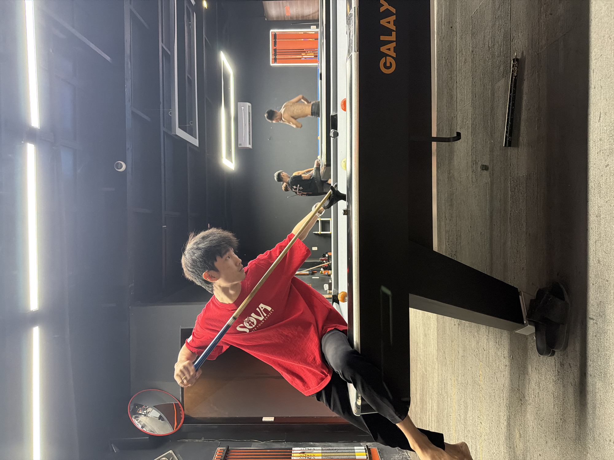 The image displays an indoor recreational facility in Đà Nẵng, Vietnam, featuring a combination of pool and bouldering activities. The main subject is a young Asian man, wearing a red T-shirt with the text "SOVA CREATE YOUR SENSES" and dark pants. He is focused on making a shot at a pool table, holding a blue and brown pool cue. He has a ring on his right hand and is barefoot, with a pair of dark sandals visible near the table's base.
In the background, two other men are actively bouldering on a dark climbing wall, which is fitted with various grips. One climber is shirtless, while the other wears a dark T-shirt. An air conditioner unit is mounted on the wall above the climbing area.
The setting is characterized by dark walls and a modern design, illuminated by several bright, rectangular LED light fixtures. A security camera is mounted high on a wall. The pool table, prominently displaying "GALAXY" in gold lettering, is situated in the foreground. Other visible objects include a round convex safety mirror and racks containing what appear to be climbing-related tools or cues, with the brand "ALLNIX" visible on some items. The overall scene depicts an energetic, multi-sport environment. The image is oriented vertically, making the standing and climbing figures appear horizontal.
