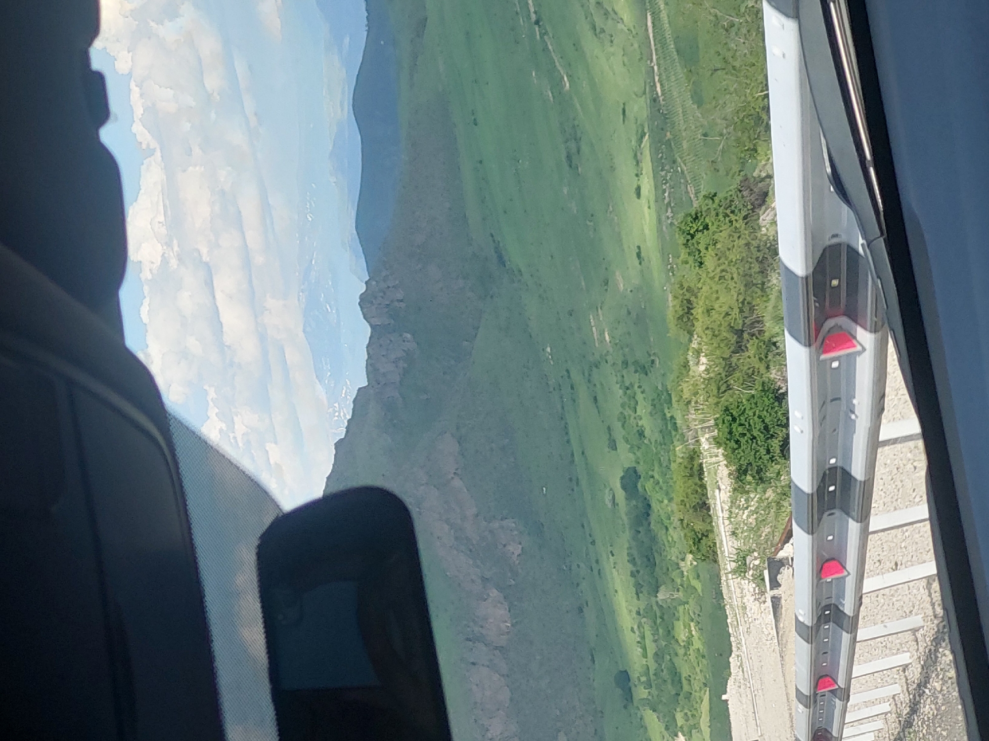 The image presents a panoramic view of a vast, mountainous landscape, likely captured from inside a vehicle traveling through Yelpin, Armenia. The scene is oriented as if rotated 90 degrees clockwise, displaying the horizon vertically.
In the foreground, elements of the vehicle frame the view. On the left, a dark dashboard, a section of the windshield, and a rearview mirror are visible. On the right, a dark vertical edge, possibly part of the car's pillar or side mirror housing, encloses the view.
The main subject is a sprawling natural environment. The sky, visible in the upper left, is bright blue with scattered white, fluffy clouds. Below the sky, layers of distant, hazy blue mountains recede into the horizon. Closer to the viewer, a prominent mountain range with rocky outcrops and abundant green foliage stands out. The landscape comprises rolling green hills and valleys, suggesting a fertile and well-vegetated area.
In the immediate foreground on the right, parallel to the vehicle, runs a distinct highway guardrail. It features a design with alternating grey and black sections and regularly spaced, reflective red markers. Beneath the guardrail, a light-colored, textured road surface or embankment is visible, along with scattered small trees and bushes.
The time of day appears to be bright daytime, under clear and sunny weather conditions. The setting suggests a journey through a scenic, mountainous region. There is no visible text in the image.