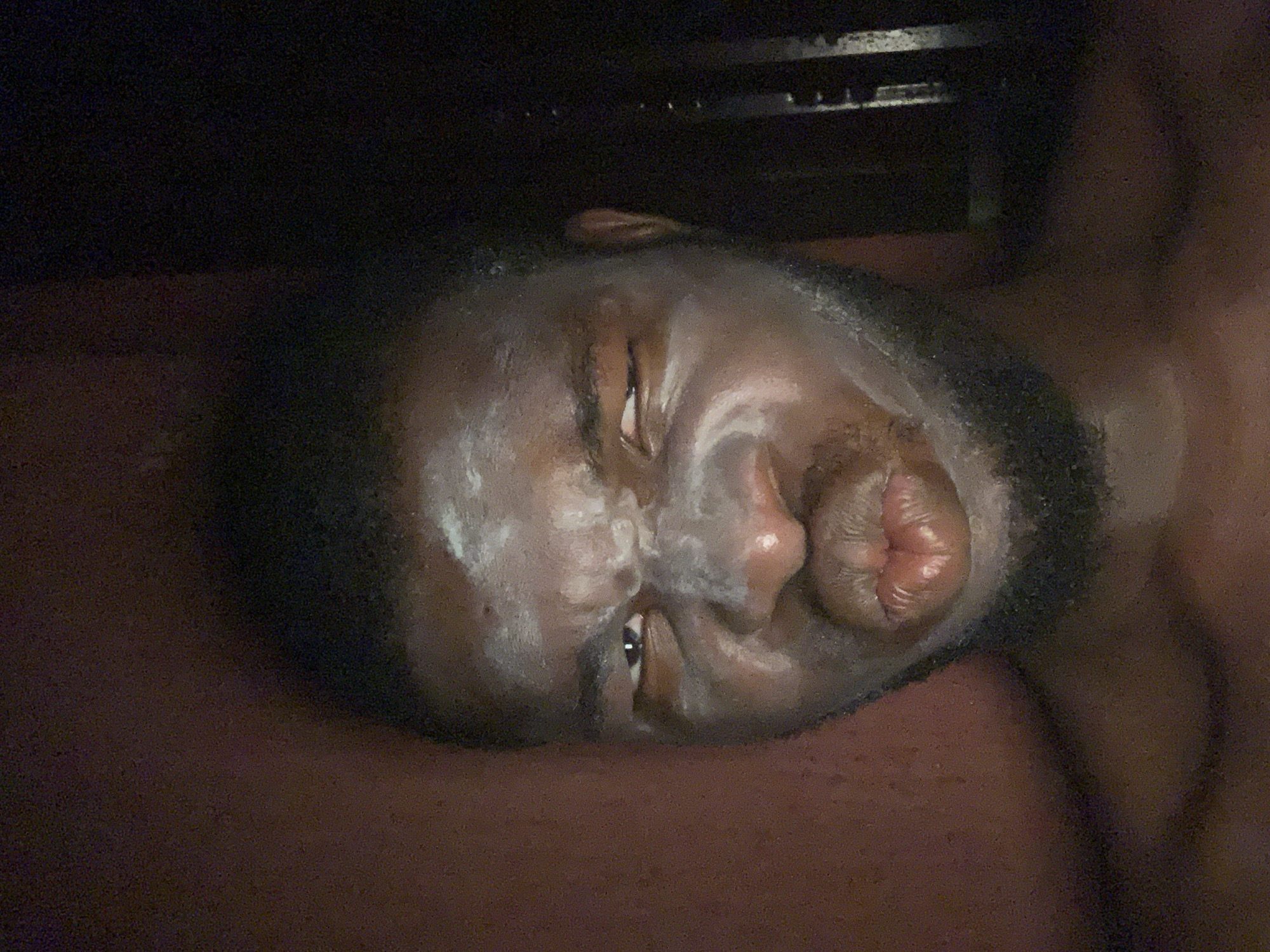 The image features a close-up, horizontal view of a person's face in a dimly lit environment, likely indoors at night. The subject appears to be a Black individual with a beard or stubble, lying down on a dark reddish-brown surface, possibly a bed or mat.
A prominent white, cream-like, or mask-like substance is visible on the person's face, covering areas around the forehead, nose, cheeks, and chin. Their eyes are partially open and squinted. The person's lips are strongly pursed and puckered, forming a distinct "fish face" or kissing gesture. The expression suggests playfulness or an intentional pose.
The background is very dark and indistinct, offering no specific details about the immediate surroundings beyond the dark surface the person is resting on. There are no other discernible objects or visible text in the frame. The overall lighting is low, highlighting the face and the white substance against the dark backdrop. The image itself does not contain any visual cues to confirm the location in Onitsha, Nigeria.