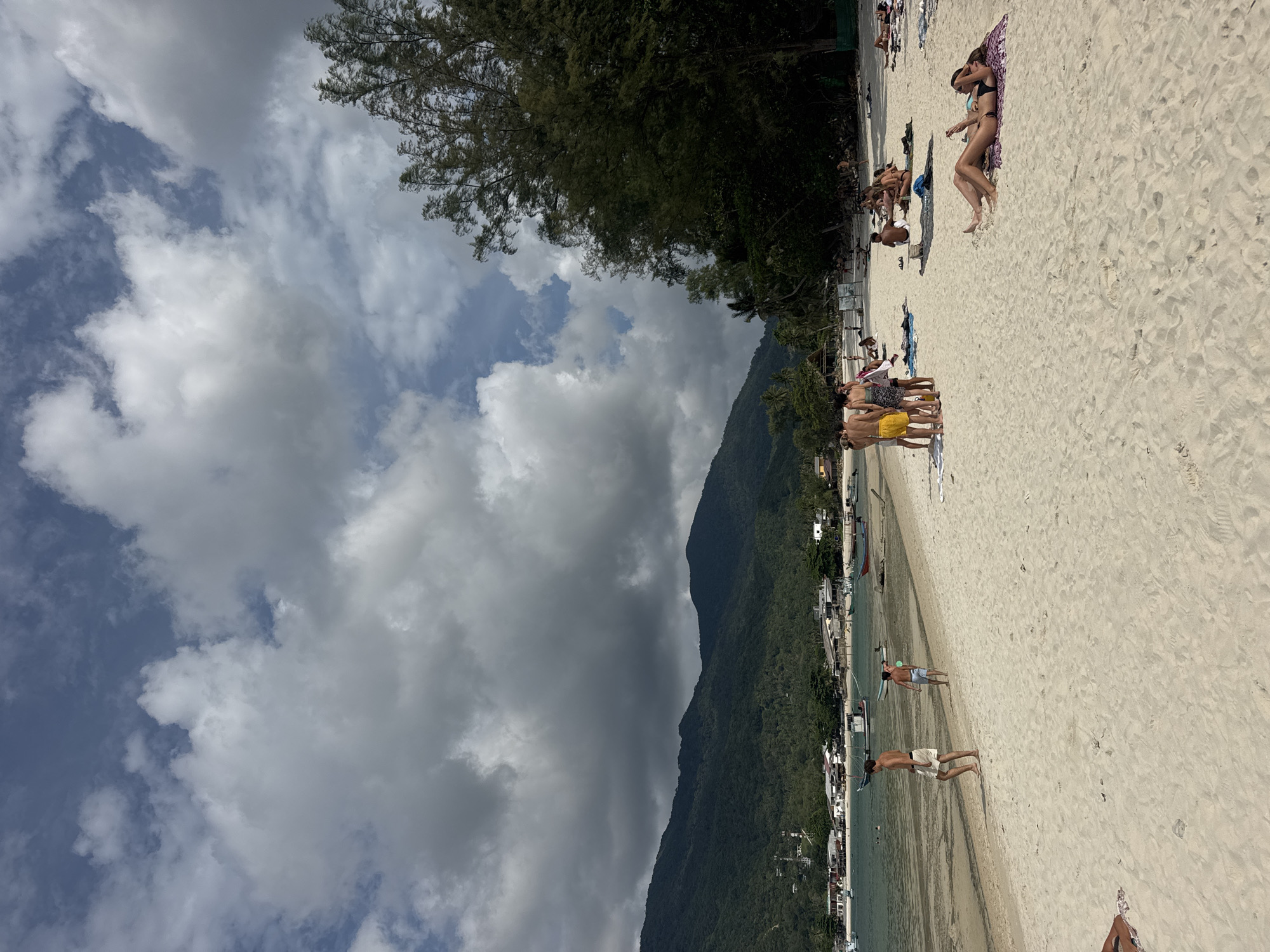 The image depicts a beach scene in Baan Chaloklum, Thailand. A mountain covered in dense green vegetation rises up from the shoreline. The sky is filled with dynamic cloud formations, including both bright white and dark grey clouds. On the sandy beach, multiple individuals are visible, engaged in activities such as sunbathing, standing in groups, and walking along the shoreline. The beach also features buildings and boats near the water's edge, suggesting a settled area.