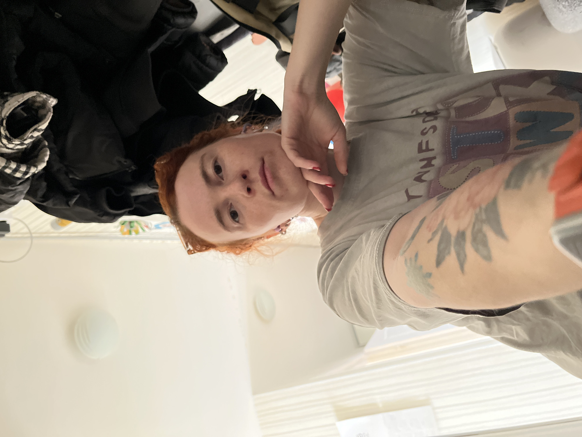 A rotated indoor selfie shows a red-haired person with fair skin, wearing a light grey t-shirt with a graphic design and a visible arm tattoo of leaves. The person has their hand near their face, with visible red nail polish. A black jacket is visible in the background on the left. The surrounding environment includes a white wall with a light fixture, a closed window, and other miscellaneous objects, suggesting a domestic setting. There is no indication of the city of Chaikovsky, Russia, in the image.