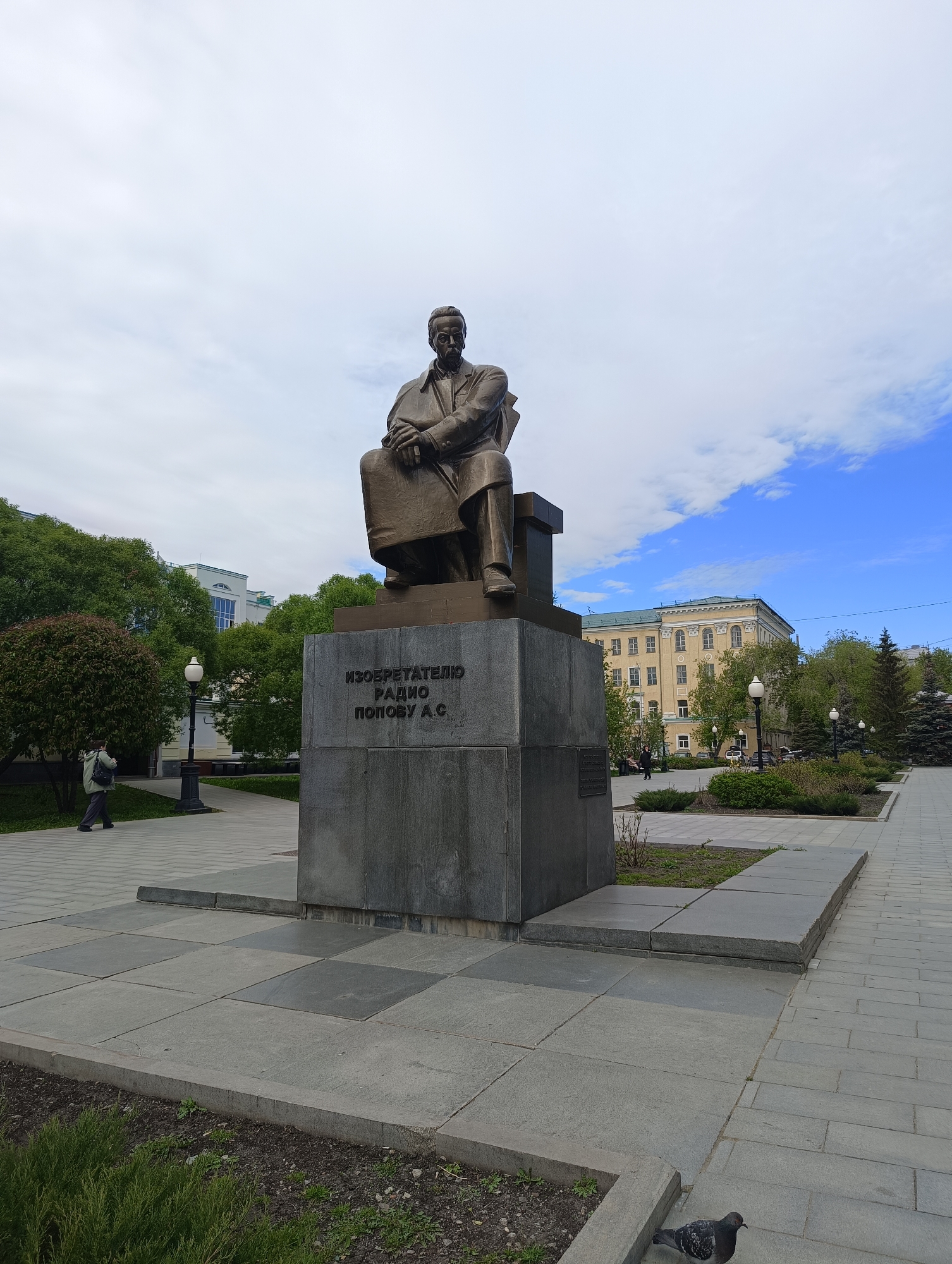 The image shows a bronze statue of Alexander Stepanovich Popov seated on a chair. The statue is mounted on a grey stone plinth with the inscription "INVENTOR RADIO POPOV A.S.". The statue is located in a public square in Yekaterinburg, Russia. The square is paved with grey stone blocks and lined with trees and lampposts. A pigeon stands on the pavement in the foreground. In the background, there is a light yellow building with a green roof. A person is walking on the paved pathway. The sky is cloudy with patches of blue.