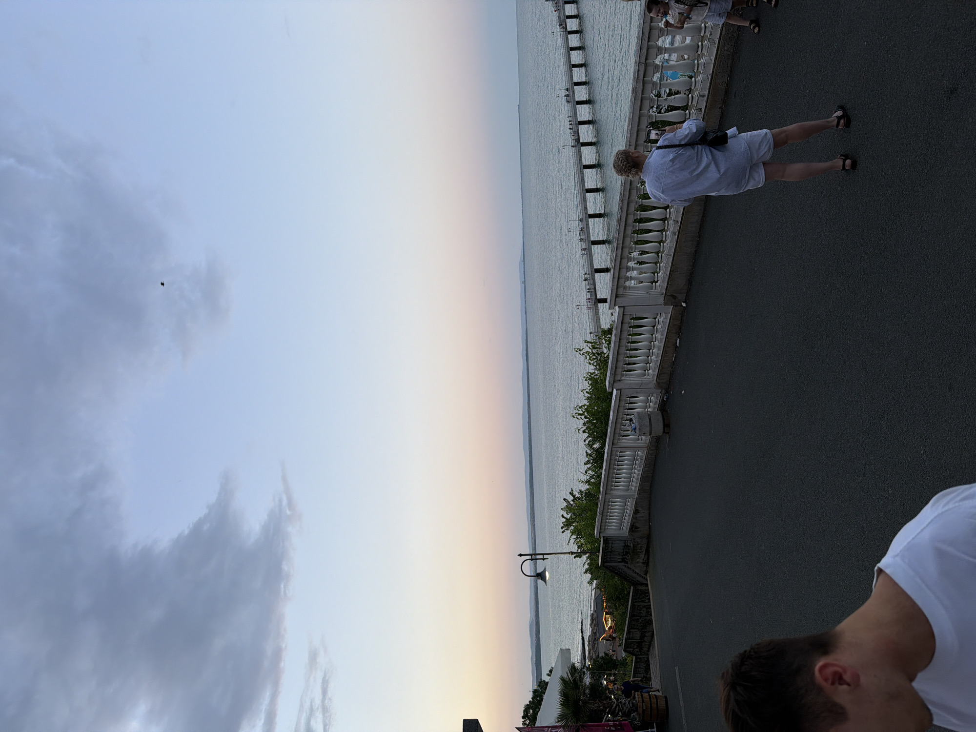 A scene unfolds on a paved walkway overlooking the sea in Burgas, Bulgaria, likely near the Sea Garden. The sky transitions from a pale blue to hues of pink and orange near the horizon, indicating either sunrise or sunset. A long pier extends into the calm, blue water, dotted with small figures walking along it. In the foreground, on the walkway, several individuals are present. A woman in a blue and white striped shirt and white shorts stands facing the sea, holding a phone and appearing to take a photo. To her right, a young child with light brown hair is also visible. Further to the left, a man in a white t-shirt is partially in frame, his back to the camera, looking down. A white balustrade runs along the edge of the walkway, providing a barrier and framing the view of the sea and the pier. Lush green foliage lines the area below the walkway. The overall atmosphere suggests a relaxed evening stroll by the coast.