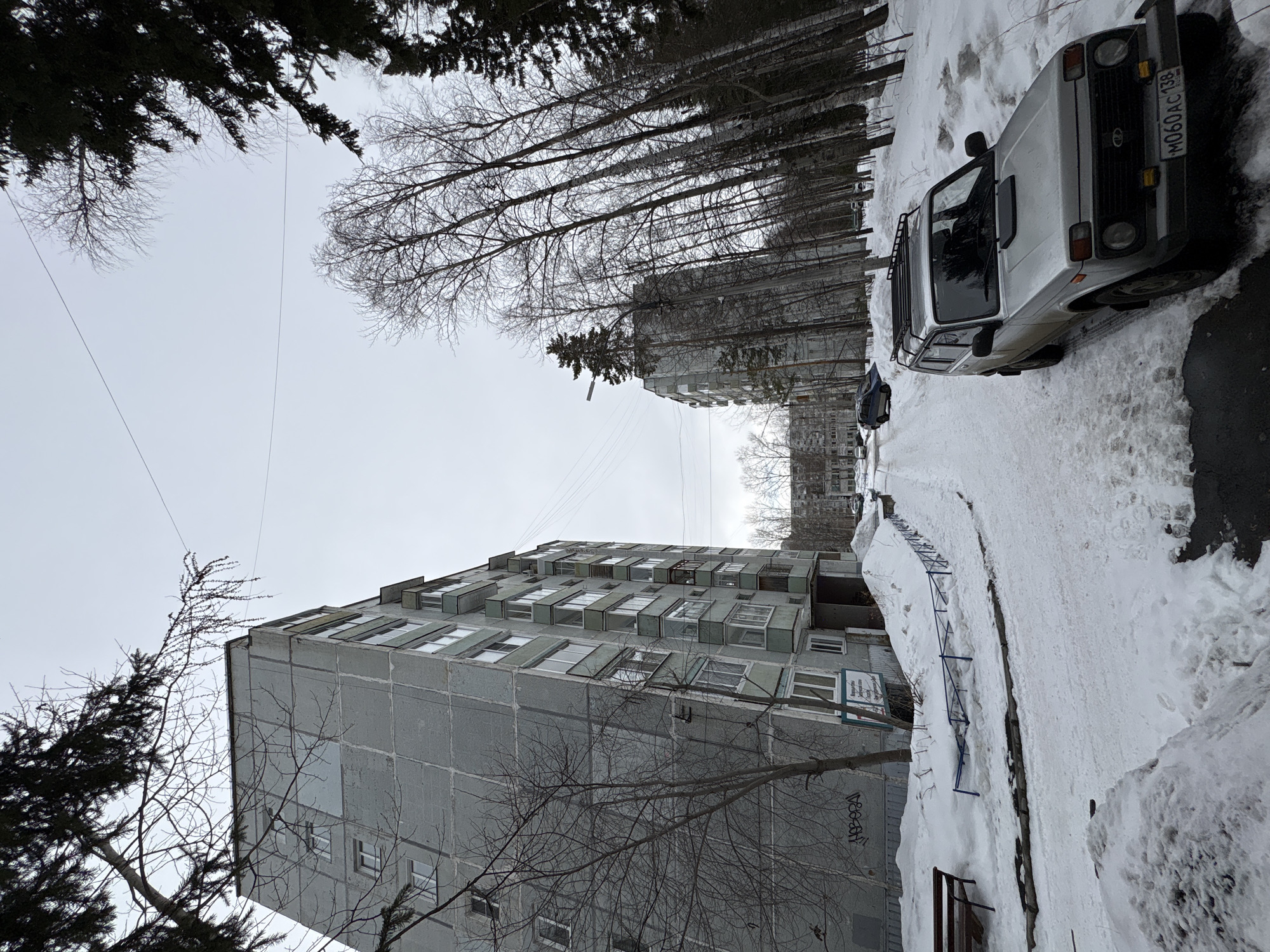 The image depicts a winter scene in what appears to be a residential area of Ust-Ilimsk, Russia. Snow covers the ground and parts of buildings. A gray, blocky apartment building with protruding window units is prominent on the left side. Bare trees are visible in front of the building. A light-colored Lada Niva automobile is parked on a snow-covered road on the right side. The license plate is visible with registration number "M060AC138". Another gray apartment building can be seen further in the background. Overhead, power lines stretch across the sky, partially obscured by tree branches. The sky is overcast, suggesting a cold winter day.