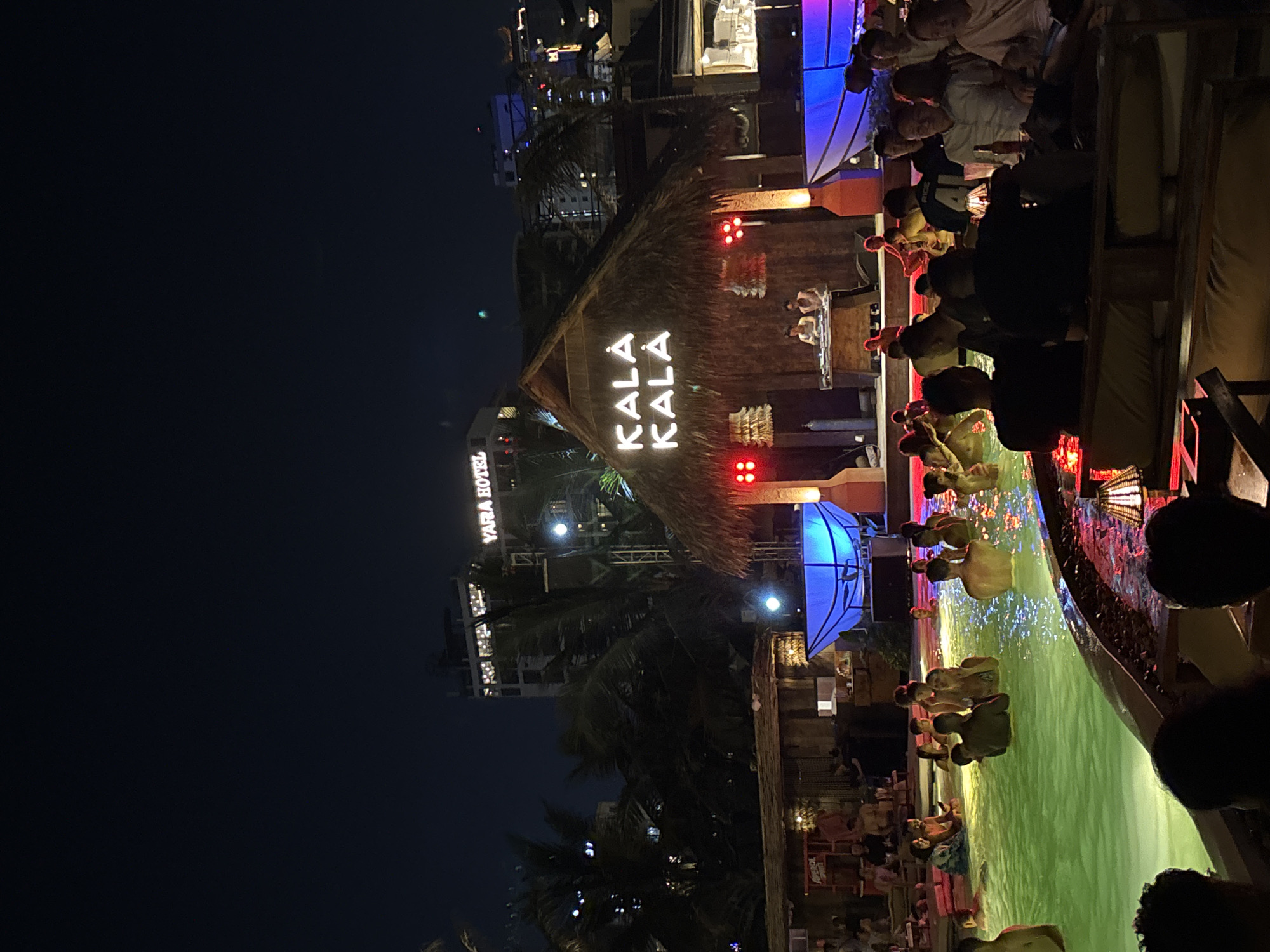 The image captures a nighttime scene in Đà Nẵng, Vietnam, featuring the Kala Kala Beach Bar. The bar's thatched roof is illuminated with a sign displaying its name. A DJ booth is visible on the structure, suggesting live music entertainment. Below the bar, a pool is visible, populated with several people swimming or wading. The pool water reflects the lights and colors of the bar and surrounding area. A crowd of onlookers is gathered, possibly patrons of the bar, watching the activities in the pool. The background includes the city skyline, with lit-up buildings and palm trees. The lighting is a mix of ambient city lights, specific lighting fixtures on the bar, and reflections on the water. The scene suggests an active nightlife with a combination of a beach bar setting and a pool party atmosphere.