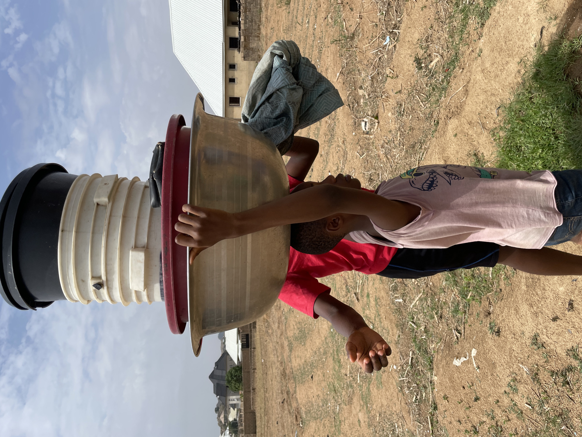 Two people wearing clothing, one in a pink shirt with skulls and the other in a red shirt, are carrying a set of large cooking containers on top of their heads. The containers consist of a black cylinder on the bottom with a white ribbed segment leading up to a red, wide, and flat container. A large tan cooking bowl sits on top of the red container. A folded grey blanket is on top of the large cooking bowl. The background shows a dry and dusty landscape with sparse vegetation, likely located in an arid climate. Some buildings are visible in the background, indicative of a residential or semi-urban area. The location is possibly Jos, Nigeria. The time of day appears to be daytime.