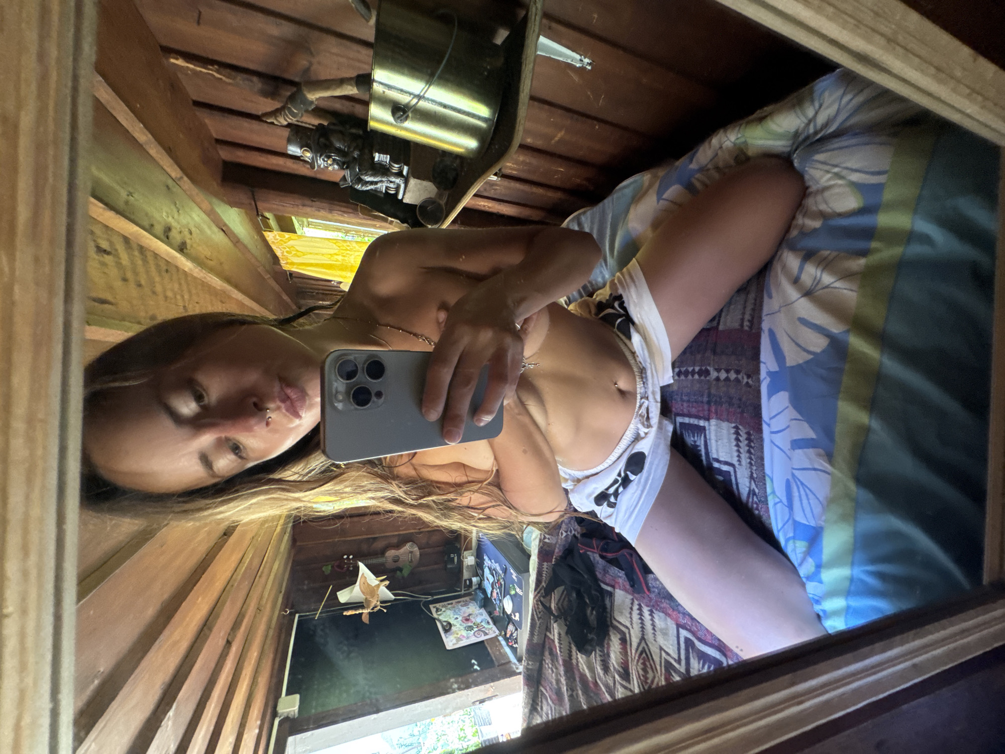A woman is kneeling on a bed, taking a selfie in a mirror. She is holding her phone in her right hand and her left hand is covering her chest. She is wearing white shorts with a black design. The bed is covered with a patterned comforter. The room appears to be made of wood, with wooden beams on the ceiling. There are various objects on shelves in the background, including a metallic bucket and a statuette.