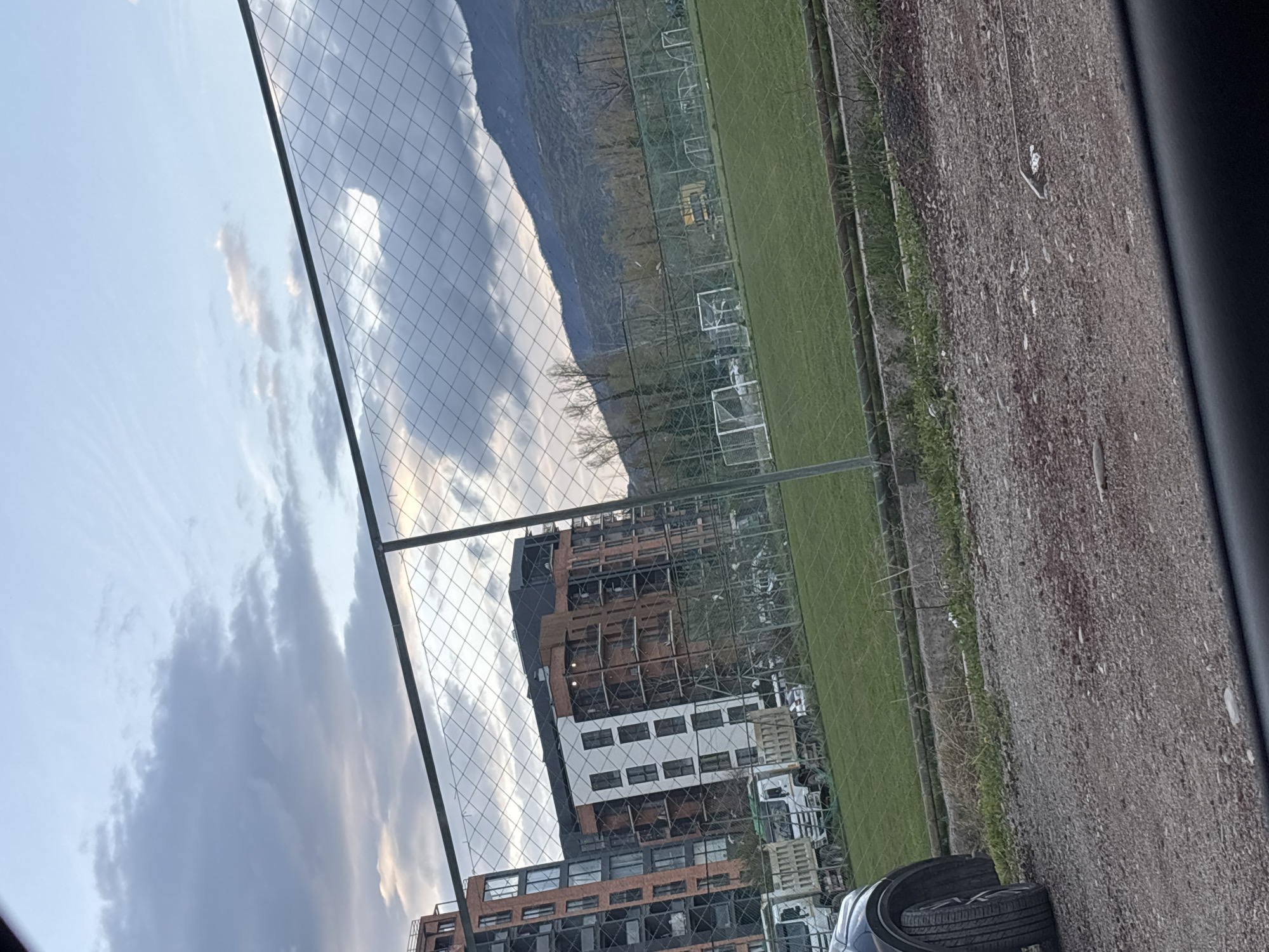 The image shows a view in Tbilisi, Georgia, likely from a vehicle looking through a chain-link fence. In the distance, there's a mountain range partially obscured by clouds. Closer to the viewer, a multi-story building under construction is visible, with a mix of exposed structure and completed sections. Next to the building is a green sports field with multiple soccer goals set up. A rough, gray, paved area occupies the foreground. Some tires are at the bottom of the view.