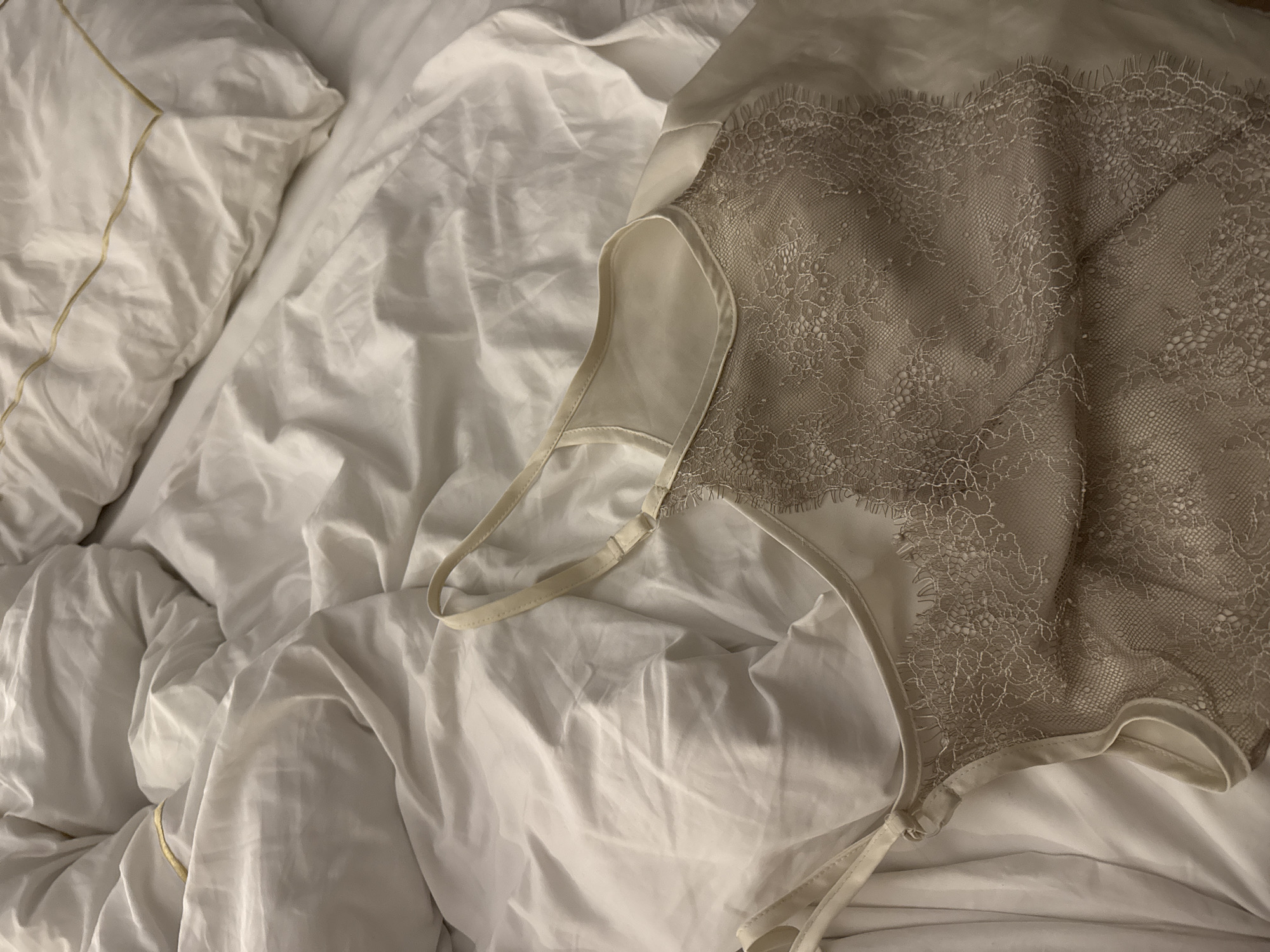 The image shows a close-up, top-down view of a beige lace camisole laid on a rumpled white duvet with subtle gold stitching on a pillow in the upper right corner. The camisole is made of a sheer lace fabric overlaid on a solid beige material, with delicate lace trim along the neckline and straps. The duvet is creased and textured, suggesting it has been used. The lighting is soft and diffused, creating a warm and intimate atmosphere. The scene appears to be indoors, likely in a bedroom. There are no people or visible actions taking place. The specific location in the city of Unknown, Egypt is not discernible from the image content.