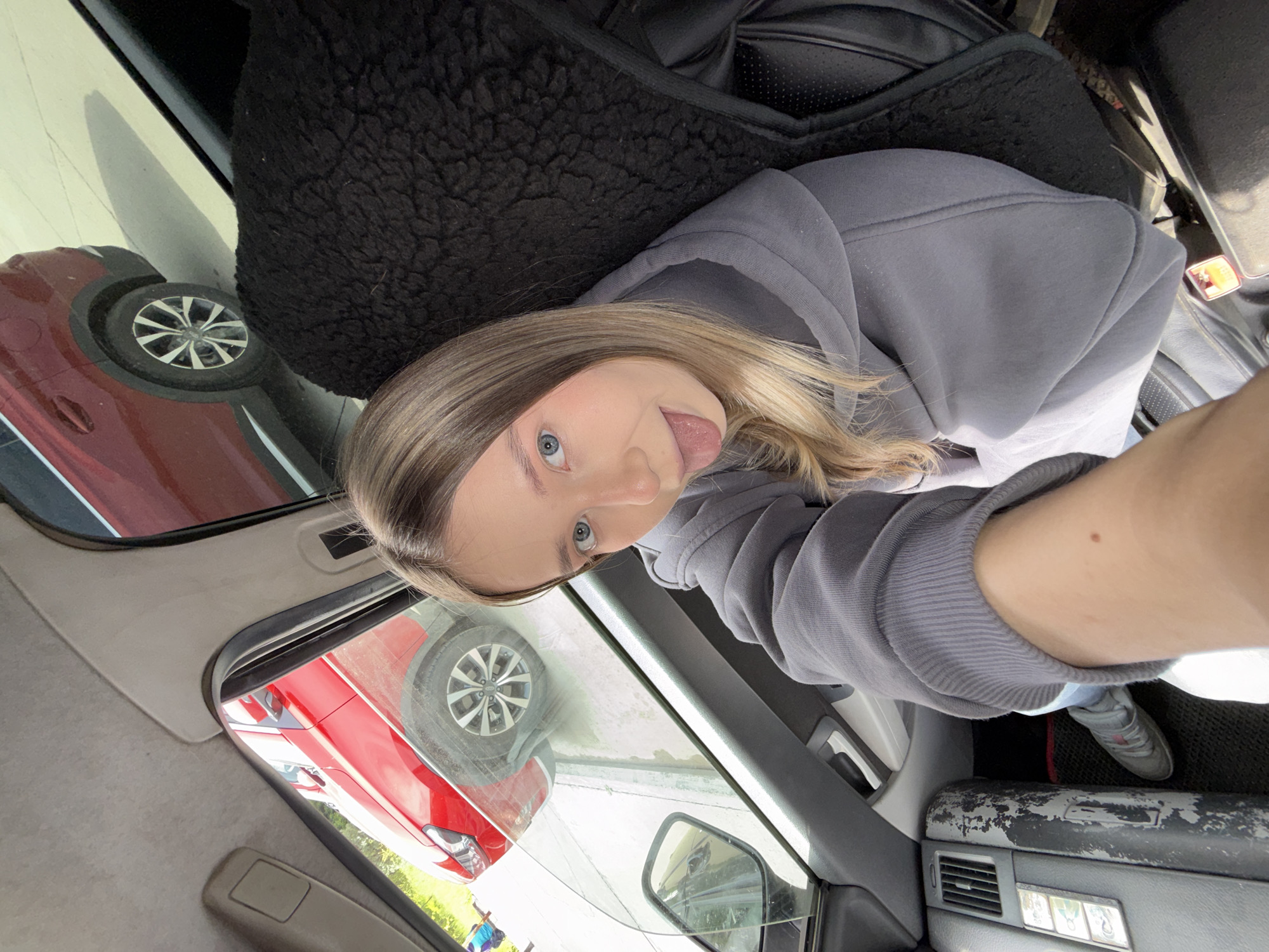 A playful selfie captures a young female inside a car, likely situated in Chaikovsky, Russia. She has light brown hair and striking blue eyes, looking towards the camera with her tongue playfully sticking out. She is dressed in a grey hooded sweatshirt.
The image is taken from a slightly upward angle within the car's interior. A black furry seat cover is visible, draped over a dark leather car seat to her right. The car's dashboard or door panel, rendered in grey, features an air vent and controls for what appear to be power windows or mirrors. A noticeable detail is the significant wear and flaking material on the lower section of the grey door panel. A grey sneaker with a white sole is visible on the dark floor mat.
Through the car window, and in reflections, a prominent red car with silver multi-spoke wheels is visible, suggesting it's parked nearby. Another red vehicle can be seen in the reflections. A glimpse of green foliage and a person in a blue top are also faintly visible outside. The bright, clear lighting indicates it is daytime, likely a sunny day. The overall atmosphere is casual and lighthearted. No legible text is visible within the frame.