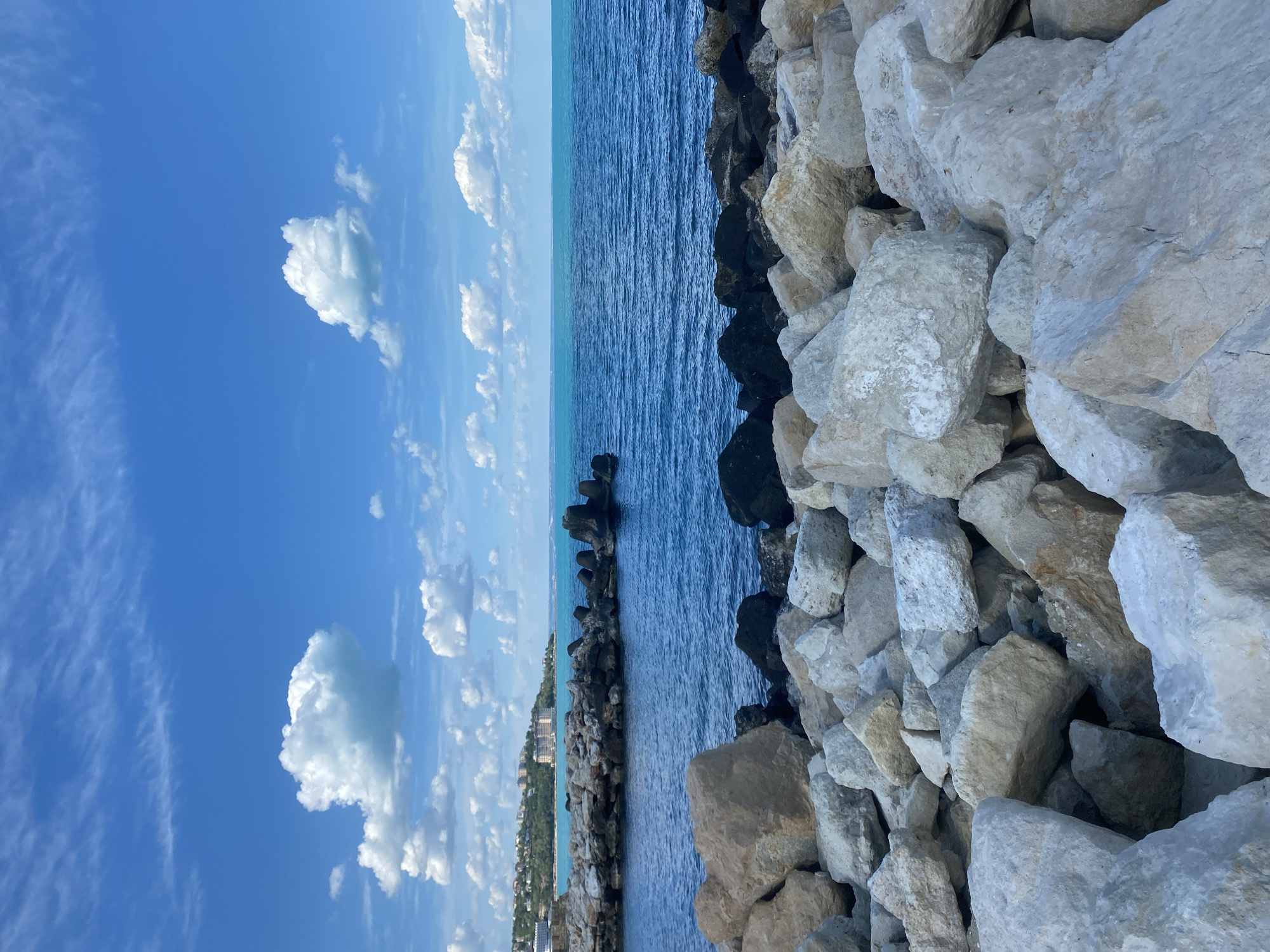 The image depicts a view from the coastal area of Varna, Bulgaria. Dominating the right side of the frame are large white rocks forming a barrier. To the left of the rocks is the blue sea, with smaller black tetrapod-shaped concrete breakwaters visible in the water. On the distant shore, a building and vegetation can be seen. The sky is a bright blue with scattered white clouds.