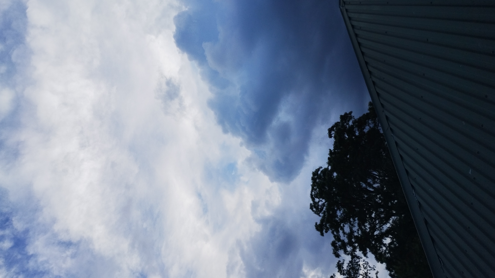 The image shows a dark, stormy sky with heavy, gray clouds. The top of a dark green, corrugated metal roof is visible in the foreground, running diagonally from the bottom left to the upper right of the frame. Behind the roof, the dark silhouette of a large tree is visible against the sky. The sky transitions from lighter, whiter clouds at the top to darker, more ominous clouds towards the bottom. There are no people or animals visible in the scene. The location is described as Unknown, Unknown, suggesting it is not a specific identifiable location.