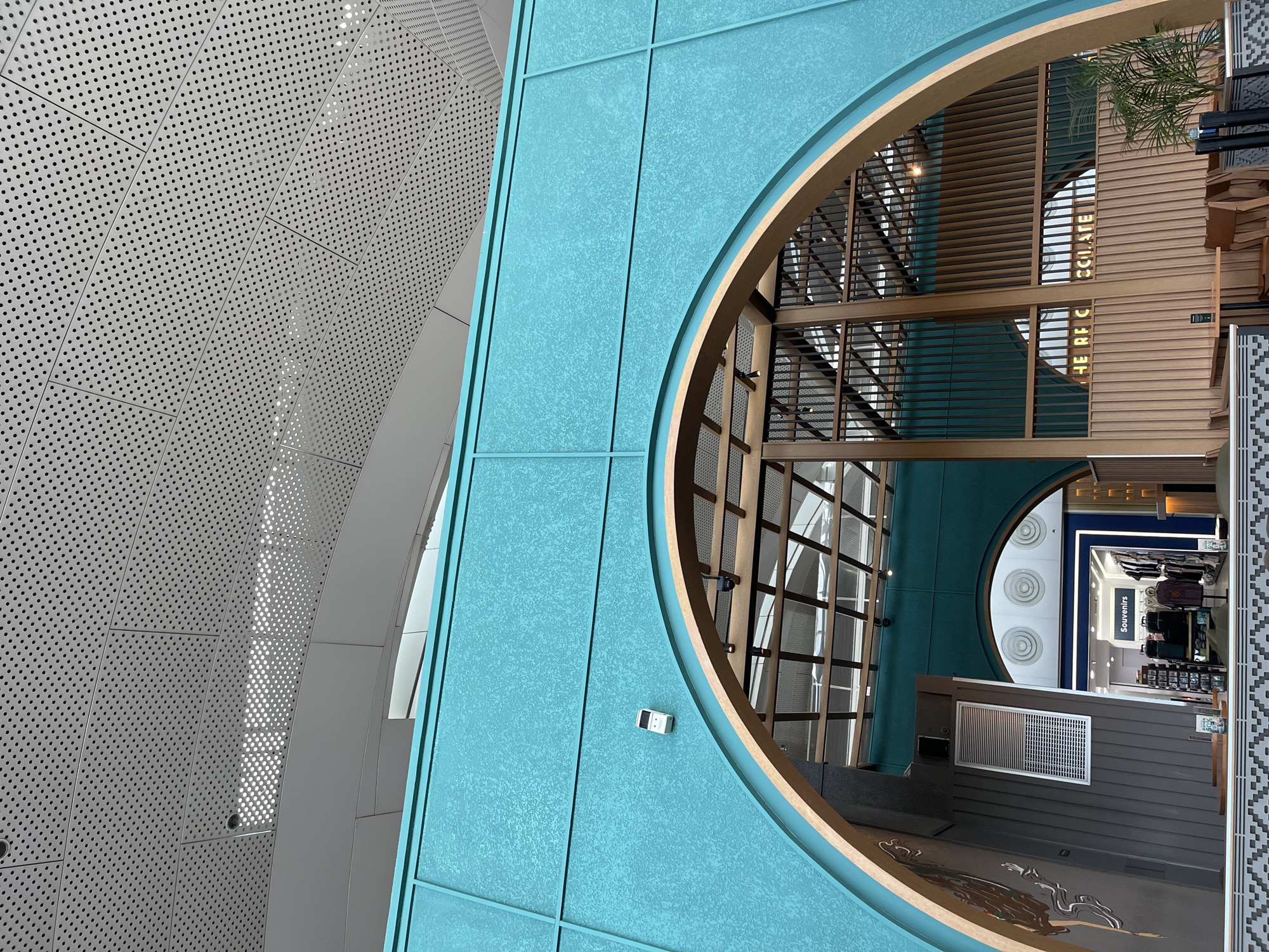This is an interior architectural photograph, likely taken in a modern building in Ramunia Dua, Indonesia. It features a view looking through a large circular opening framed in wood and teal-colored panels.
To the left is a perforated white metal wall. The area beyond the circular opening shows a multilevel space with wooden structures, and storefronts with merchandise labeled "Souvenirs". The lighting appears to be a mix of natural and artificial.