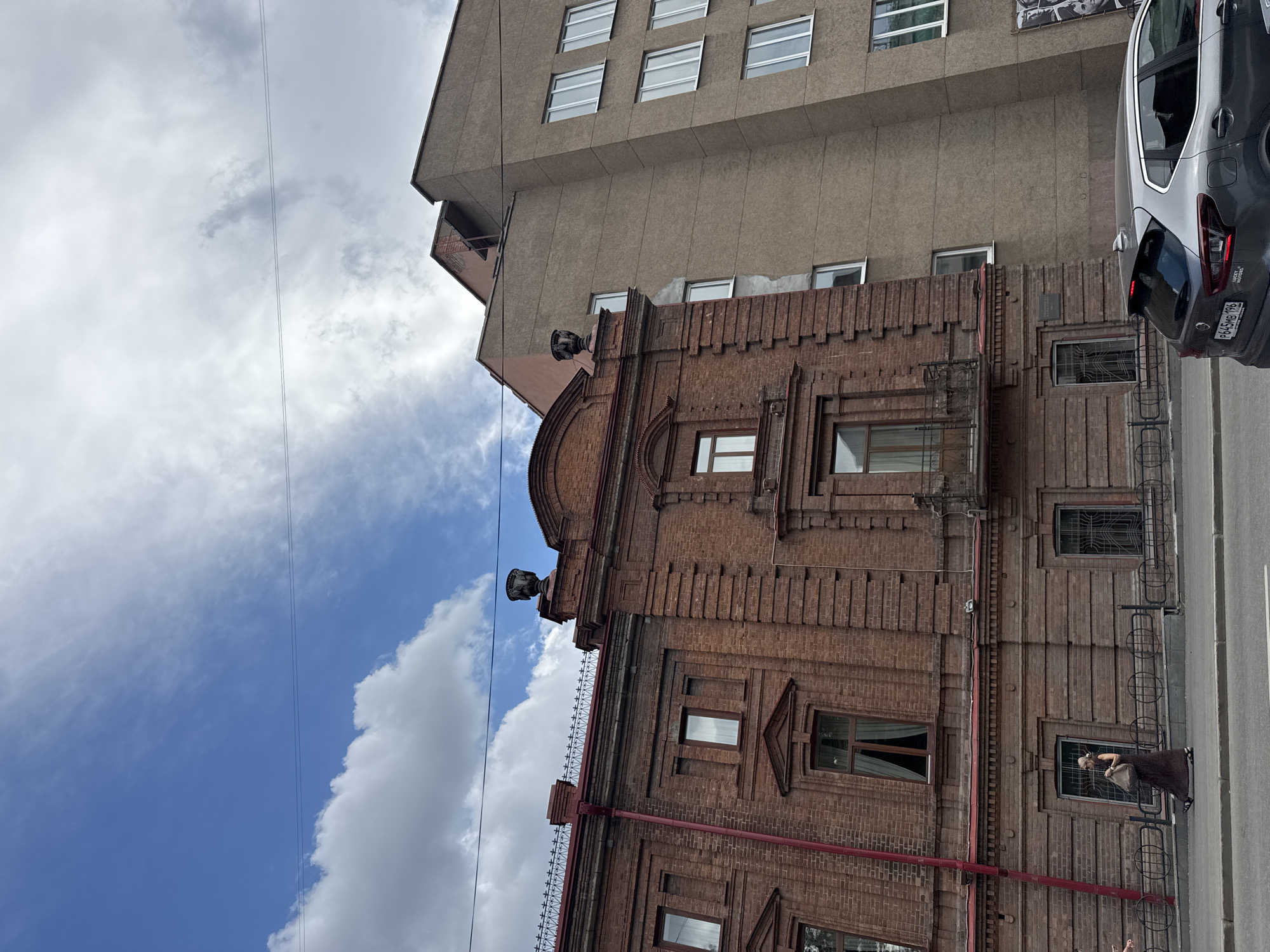 This media file captures a street-level view of a multi-story building, likely in an urban environment given the presence of a car and building structures. The building exhibits distinct architectural styles; the lower section is constructed of dark red brick with ornate detailing around the windows and a decorative cornice. Above this, a more modern section of the building rises, characterized by a plain, beige-toned facade with multiple rows of rectangular windows.
The sky is visible in the background, featuring a mix of bright blue sky and fluffy white clouds, suggesting it is daytime and the weather is fair. Electrical wires are strung across the frame, a common sight in urban settings. A dark-colored car is parked on the right side of the image, partially obscuring the view of the building's facade. One person is visible on a balcony or ledge of the brick building, looking out. The scene is static, with no specific activity or event clearly depicted beyond the presence of the car and the person. The overall impression is one of an ordinary day in a city, with the contrast between older and newer architecture being a prominent feature. The image is oriented vertically.