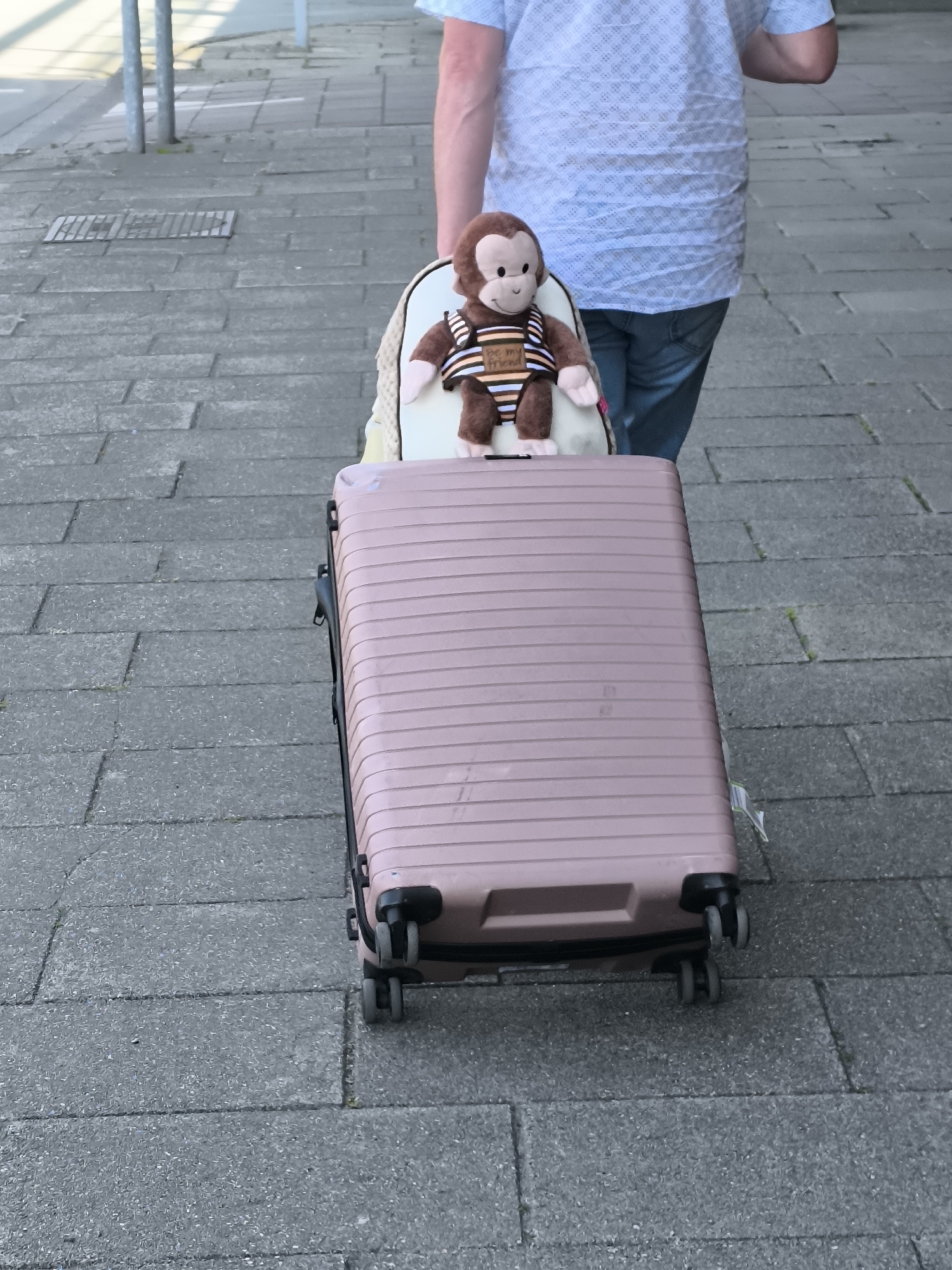 A man is seen from behind, pulling a rose-colored suitcase. A small white backpack is strapped on top of the suitcase, and a plush monkey wearing a striped shirt is placed on top of the backpack. The setting appears to be a paved area in Hoofddorp, Netherlands. The walkway consists of grey, rectangular paving stones with narrow gaps between them. In the background, there are vertical metal posts near a street. The man is wearing a light blue patterned shirt and blue jeans.