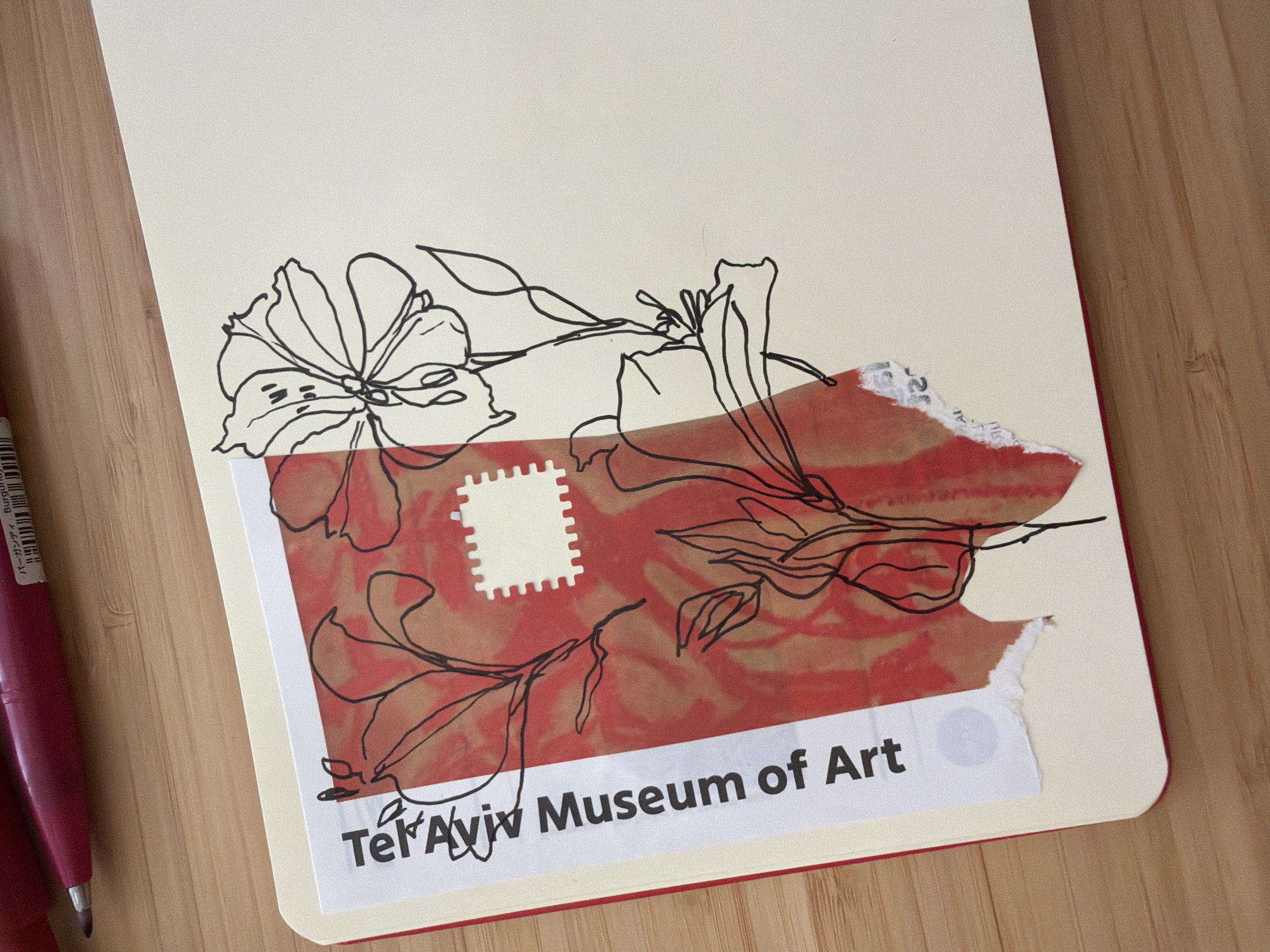 A close-up image shows a notebook on a wooden surface. The notebook has a cream-colored page with a red border visible along the bottom edge. A black line drawing of a flower and foliage is on the page. A torn piece of paper with a red marbled pattern and a white stamp-shaped cutout covers a portion of the drawing. The phrase "Tel Aviv Museum of Art" is printed on a white strip adhered to the bottom. A burgundy-colored pen is visible on the left side. The location in the city of Bat Yam, Israel is not provided in the image.