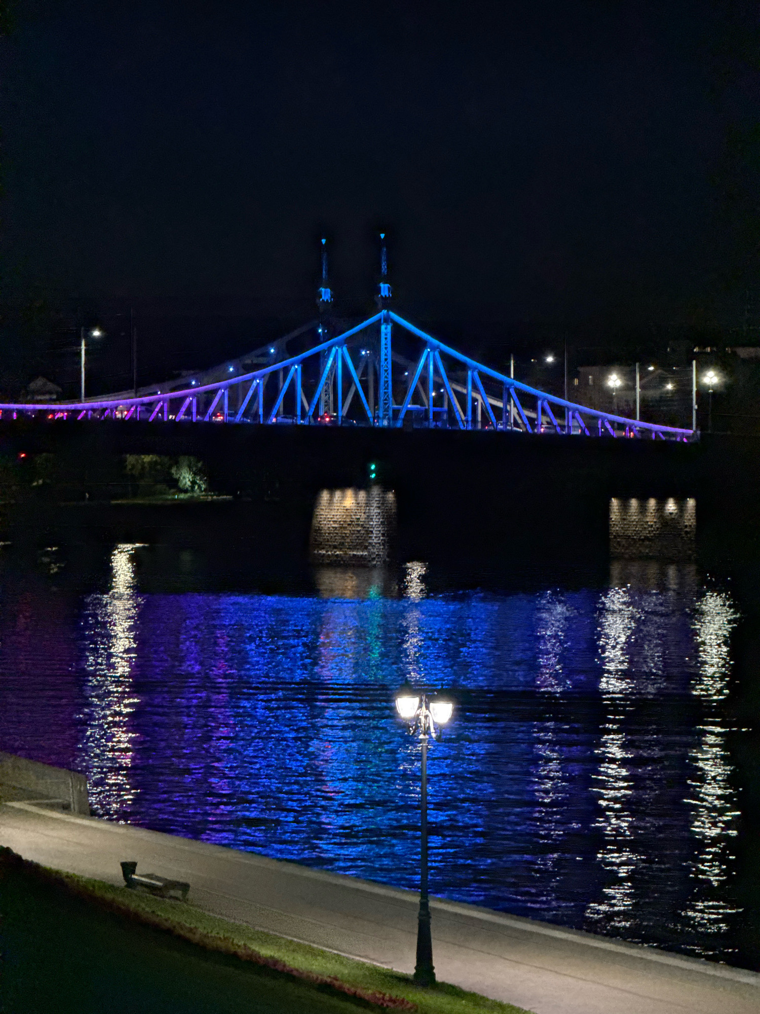 This is a nighttime photograph of the Starovolzhsky Bridge in Tver, Russia. The bridge, illuminated with blue and purple lights, spans across the Volga River. The lights from the bridge cast colorful reflections on the dark water. On the riverbank in the foreground, a pathway is visible with a lamppost and a bench. The overall scene depicts a well-lit urban infrastructure at night, highlighting the bridge and its illuminated features against the dark sky and water.