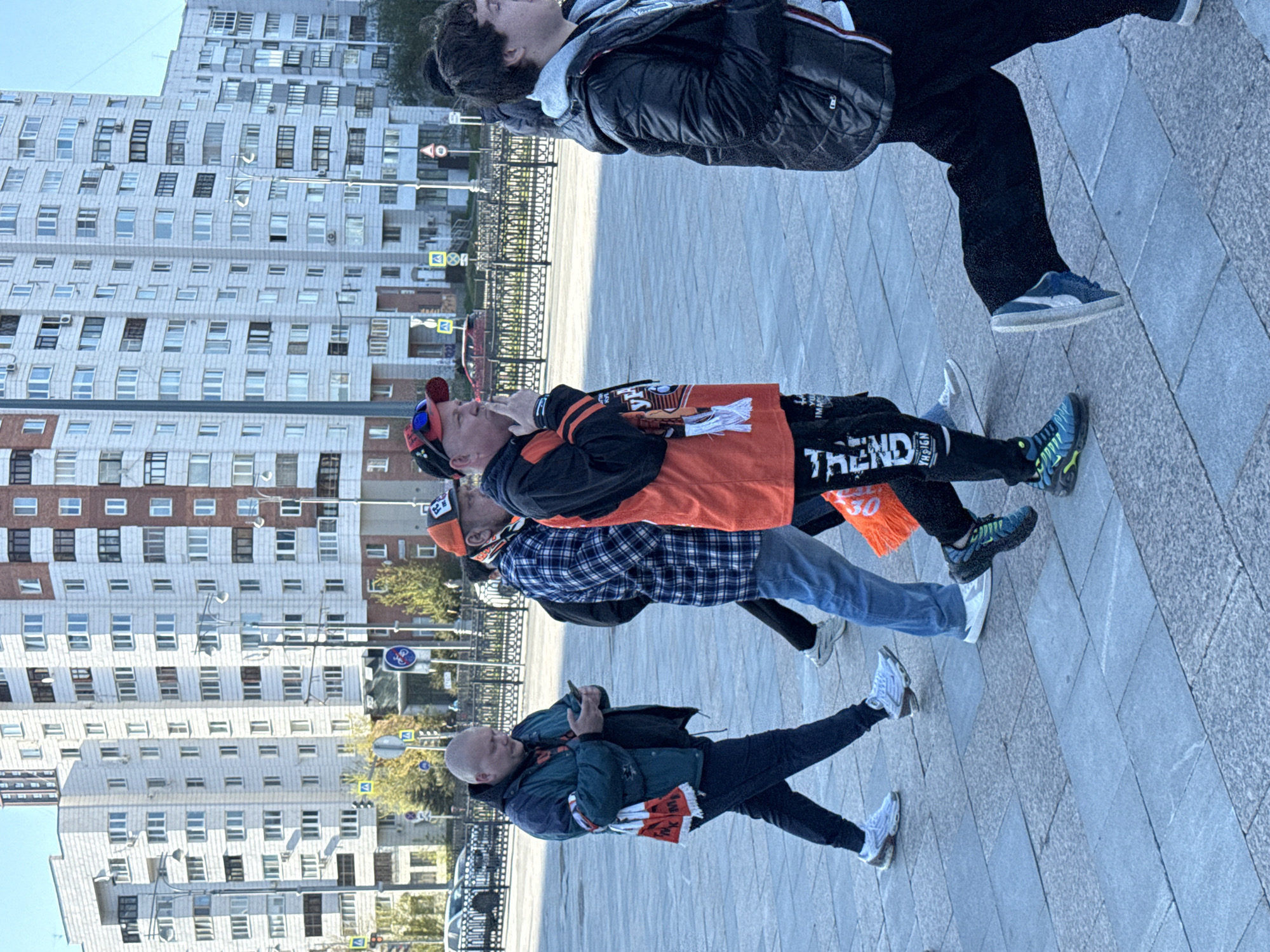 The image captures a group of four people on a paved area, seemingly in Yekaterinburg, Russia, based on the urban landscape. In the background, a tall, predominantly white building with numerous windows is visible. Starting from the bottom, the first individual is a bald man looking at a cellphone wearing a dark jacket with an orange and white scarf. The second person is wearing a plaid shirt and jeans. The third person is wearing an orange scarf and black pants with the word "TREND" on them. The fourth person is wearing black and is looking up. The people appear to be moving, possibly walking together. The pavement is made of large, rectangular tiles. In the background, there is a black metal fence and various traffic signs. The weather appears to be clear and bright, judging by the sky and the lighting conditions.