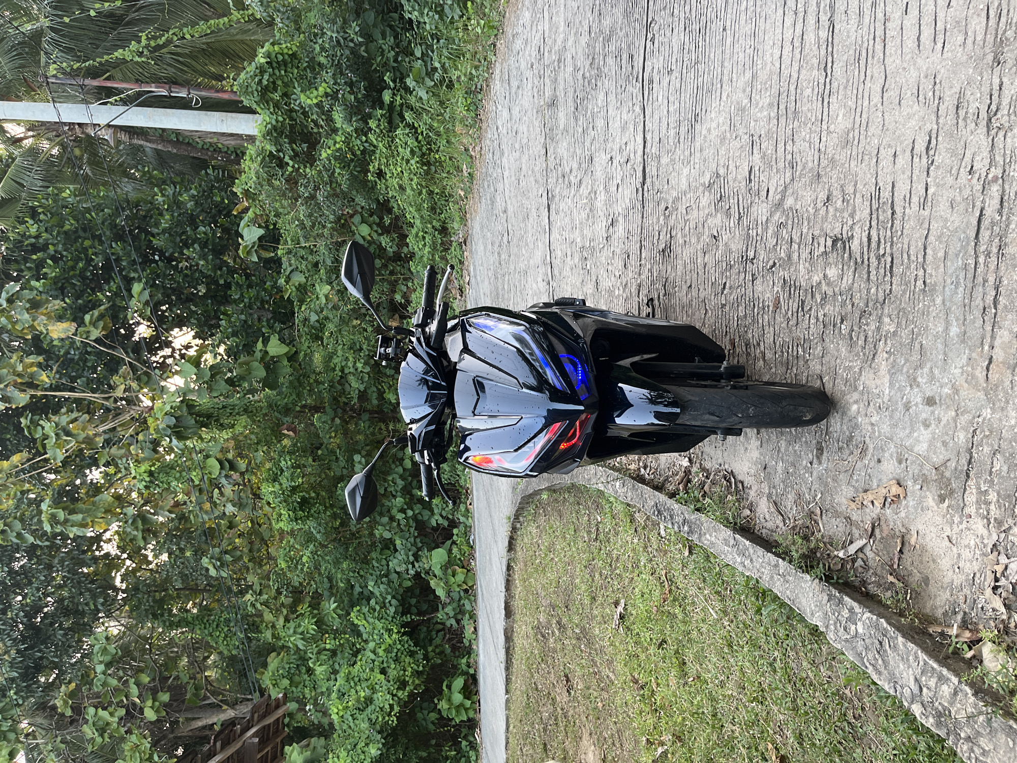 A black motorcycle is parked on a concrete path with a grassy area to the left, in Legonipah, Indonesia. The motorcycle is facing the camera and has its headlights on, emitting red and blue light. Lush green trees and vegetation surround the scene. A utility pole and wires are visible in the background. The ground is made of rough concrete with visible cracks and debris.