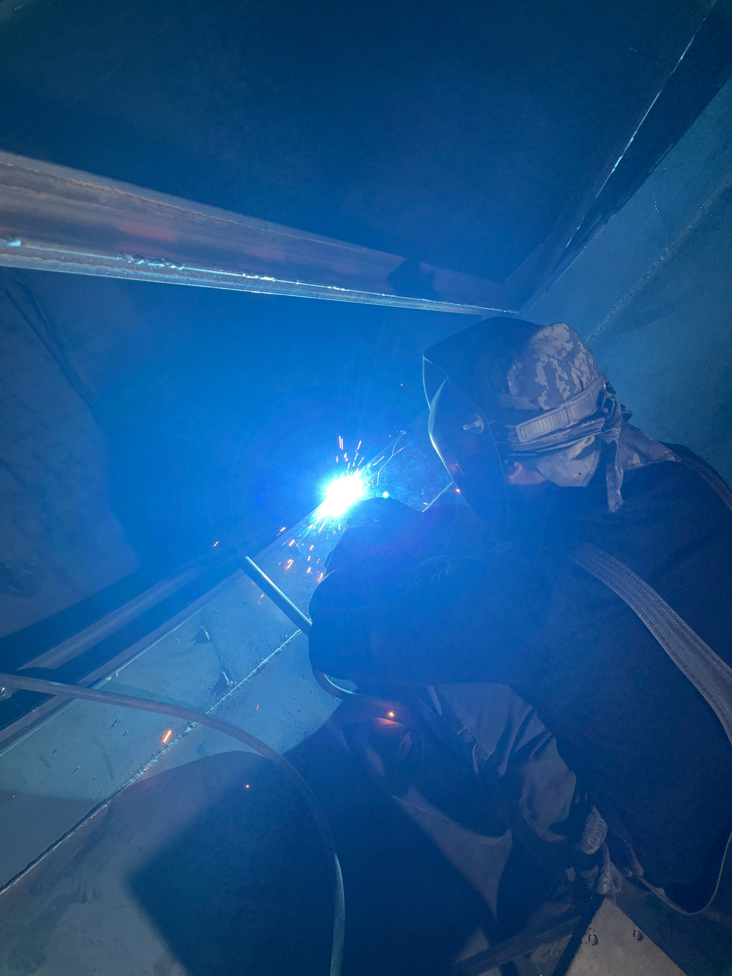 The image depicts a welder working in an industrial setting, possibly a workshop or construction site, in Taganrog City District, Russia. The welder, wearing protective gear including a helmet and a patterned bandana, is in the process of welding a metal structure. Sparks are visible, emanating from the welding arc which is intensely bright and blue. The environment appears enclosed, with metal beams and surfaces visible, reflecting the blue light from the welding. The overall scene is dimly lit, emphasizing the brilliance of the welding process.